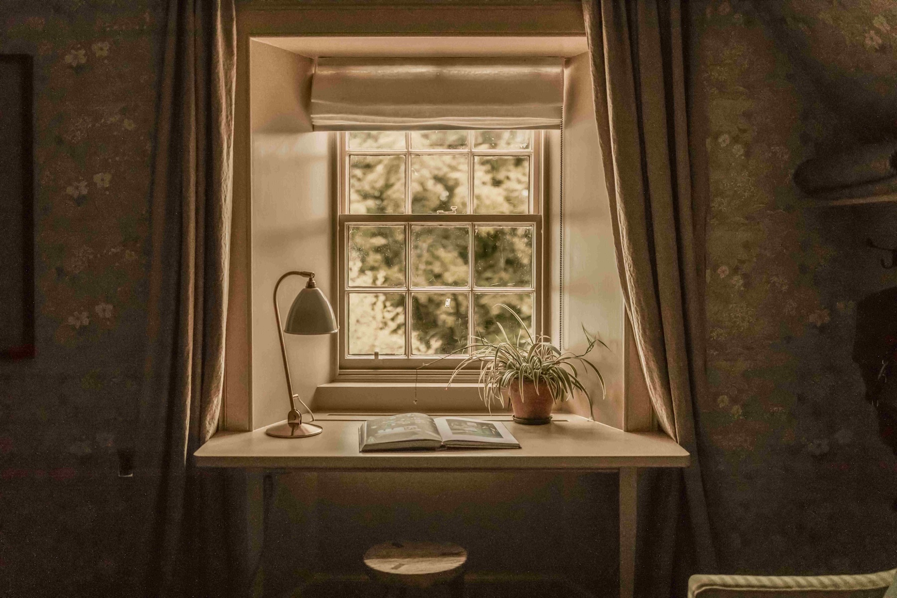 A vintage Bestlite desk lamp, open book and spider plant on a window-seat desk set into floral wallpaper, framed by linen curtains and a sash window, in a Scottish Highland guesthouse.