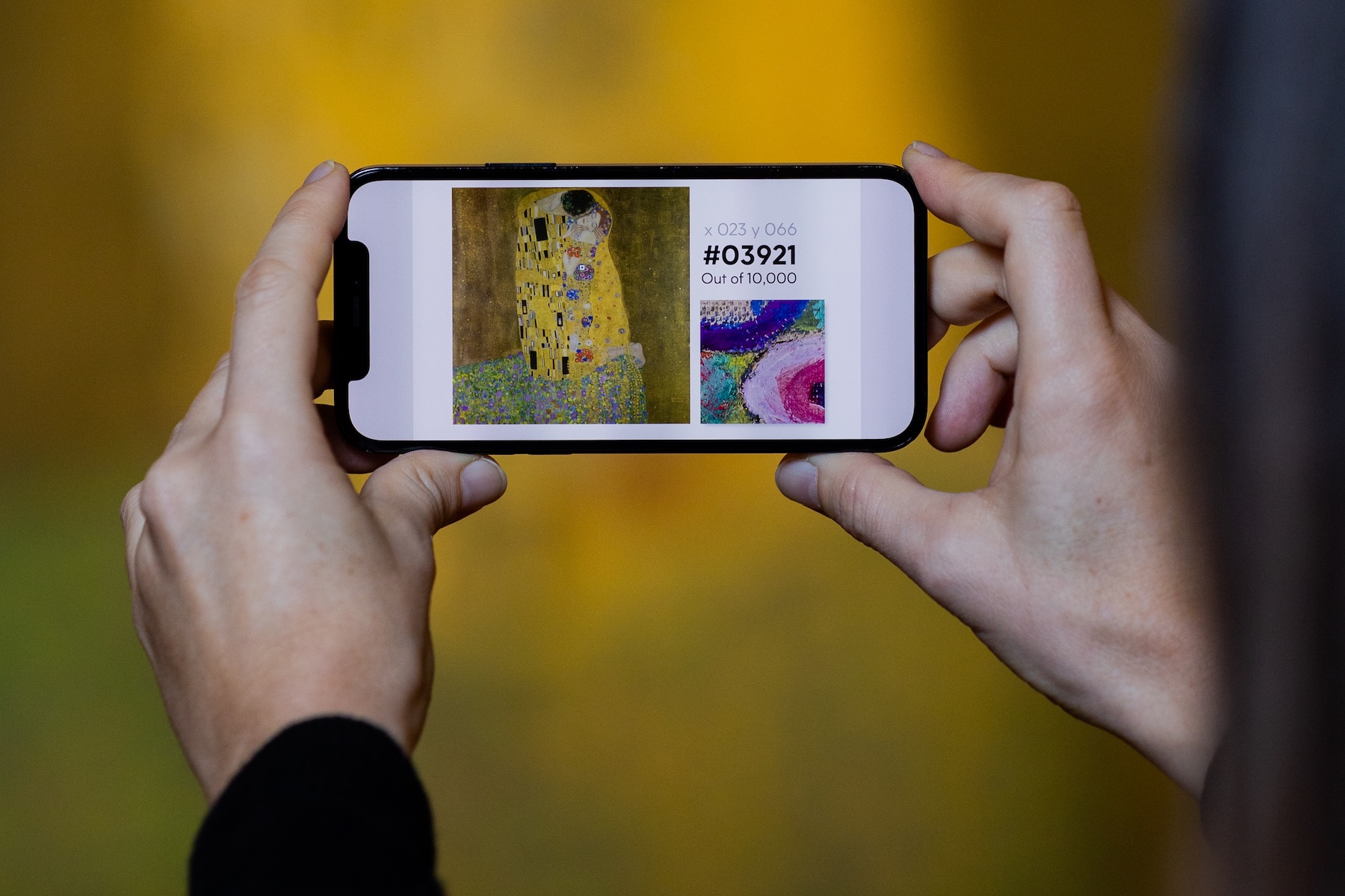 Hands holding a smartphone showing Gustav Klimt's The Kiss as a numbered digital collectible, number 03921 of 10,000, against a gold background