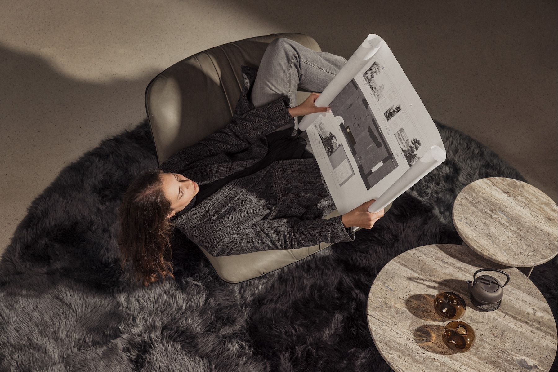 Overhead view of a woman reclining in a grey leather lounge chair, studying an Egetemeier architectural floor plan on a dark sheepskin rug, with travertine side tables and a cast-iron teapot.