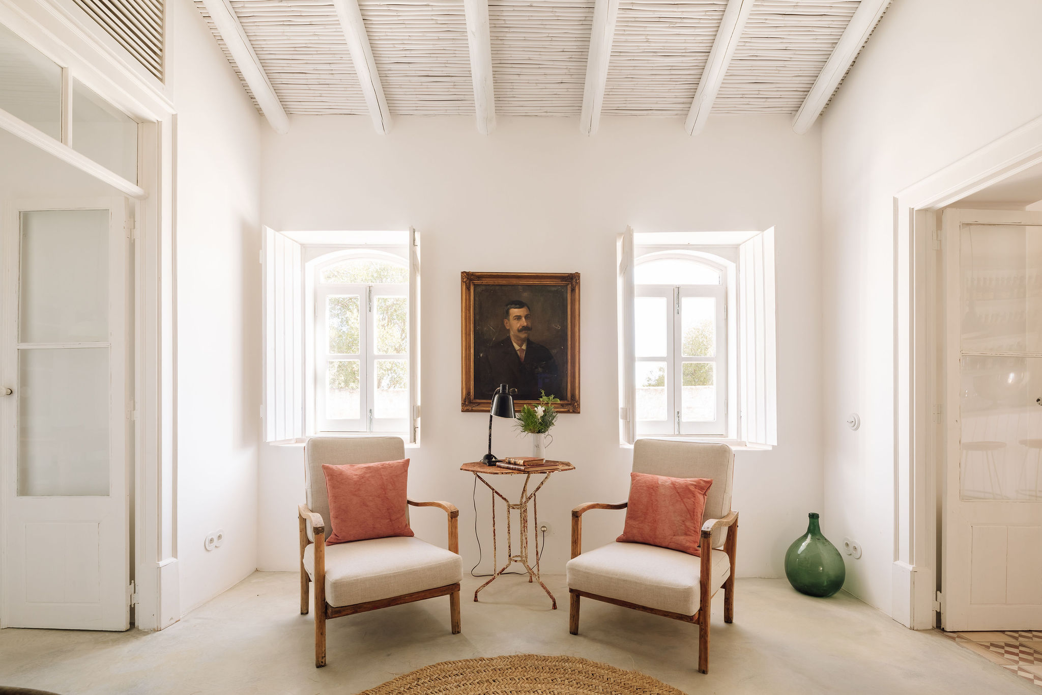 A whitewashed sitting room at Hospedaria in the Algarve, Portugal, with beamed ceiling, two linen armchairs in terracotta and an antique portrait between the shutters.