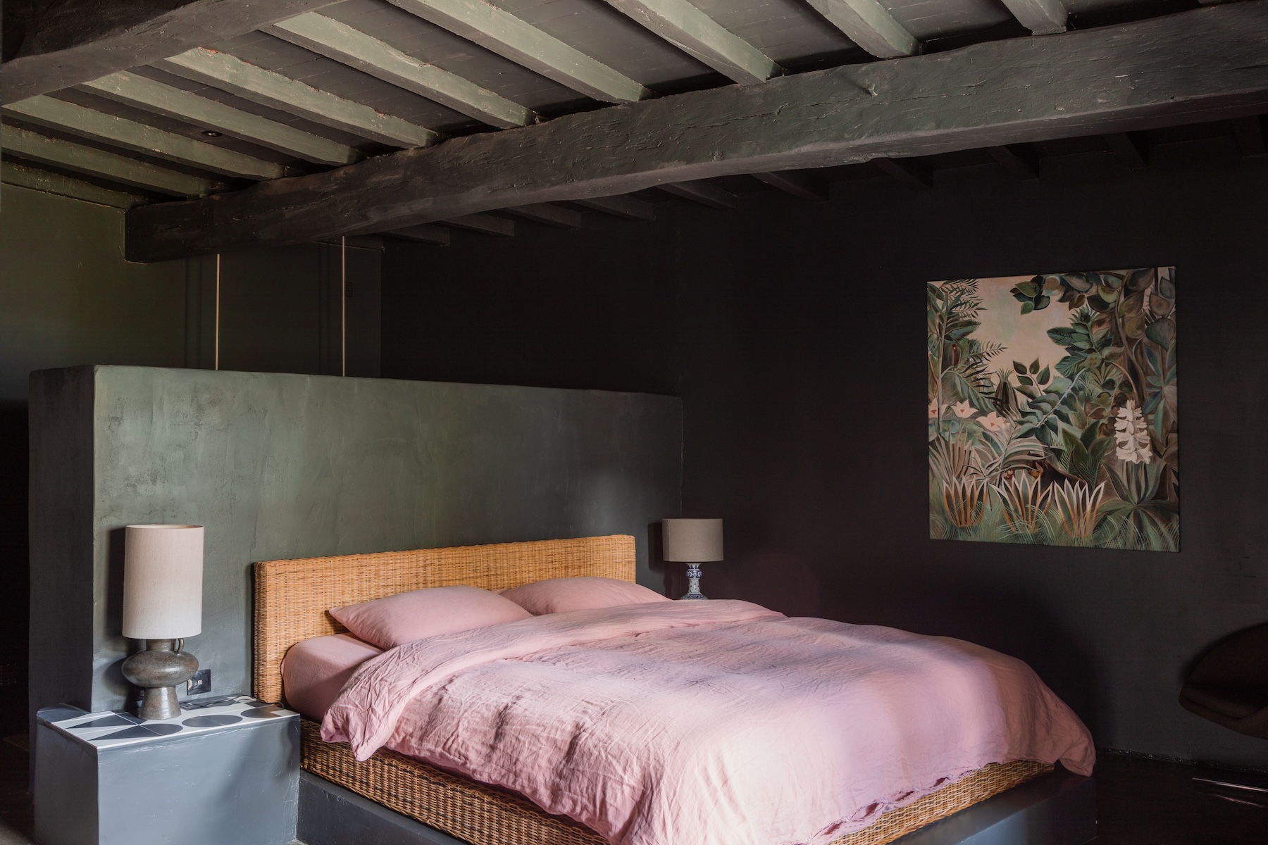 A dark-walled bedroom at Maison Duroy, Gascogne: a rattan bed, pink linen and a jungle painting beneath painted beams.