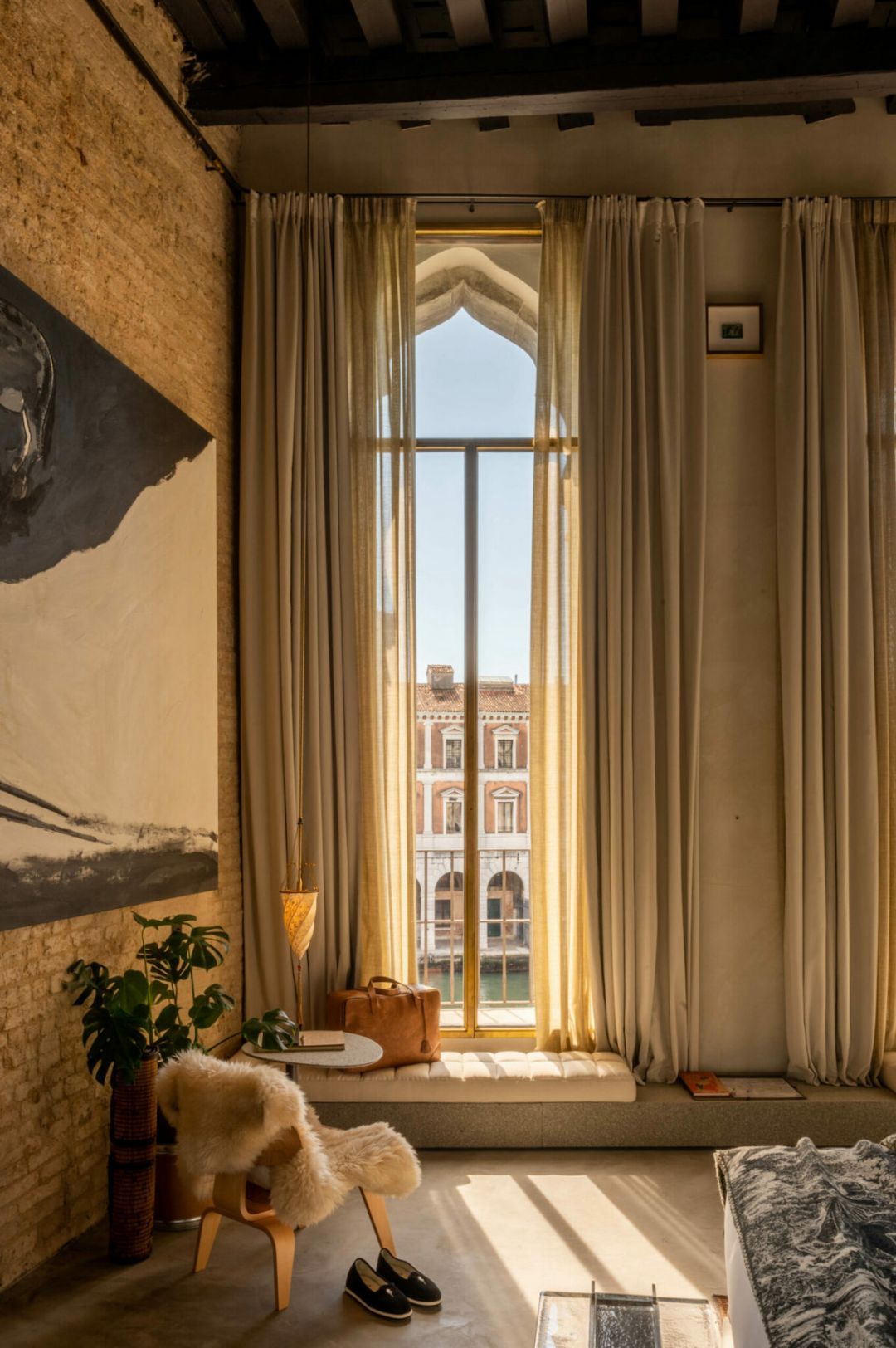 A tall Gothic window framing a Grand Canal view in a guest room at Venice M'Art, Venice, with sheer curtains, a cushioned window seat and a sheepskin-draped chair.