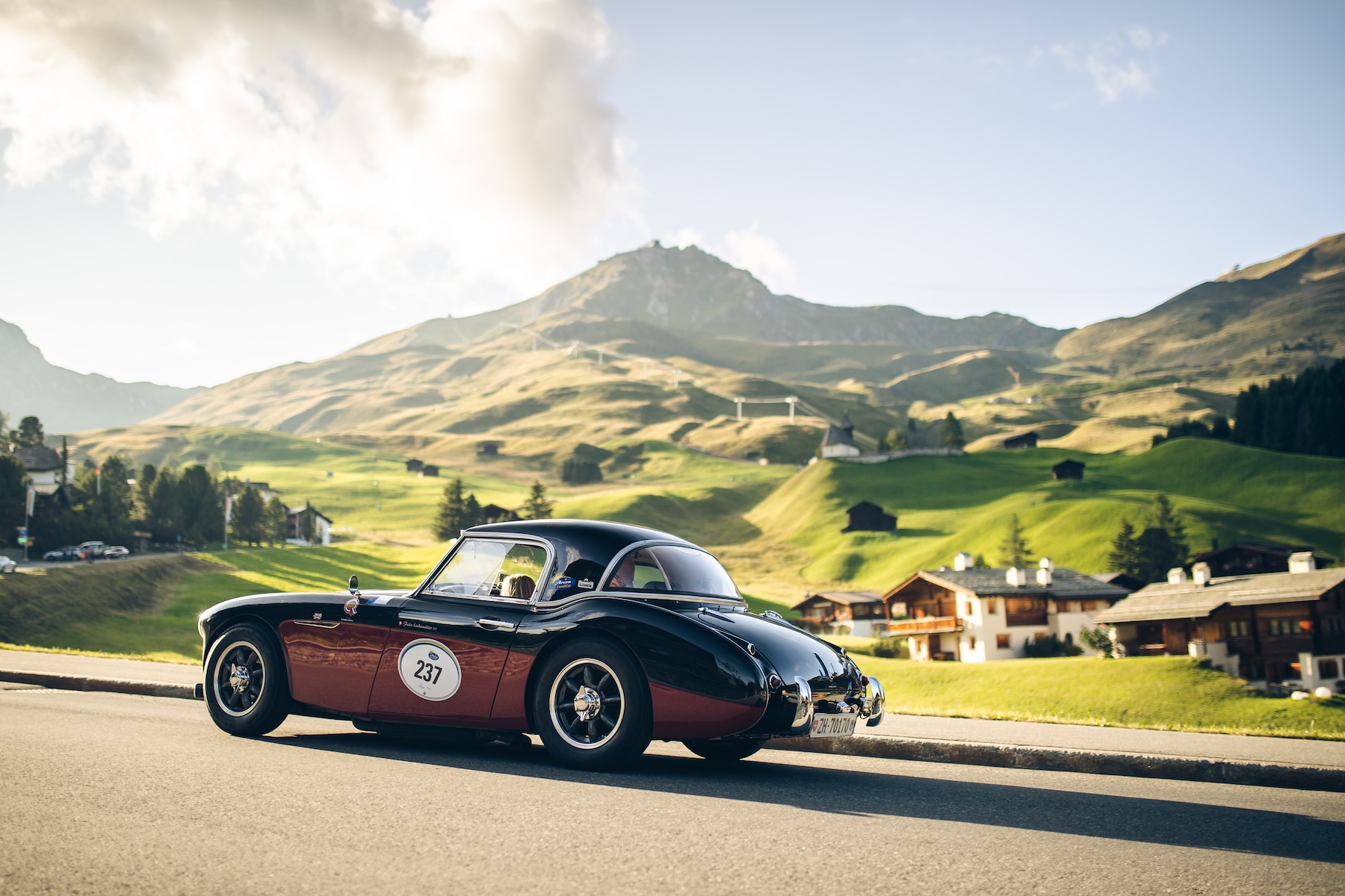 Two-tone black and maroon classic British sports car, number 237, on the road above Arosa with chalets and peaks behind | The Aficionados