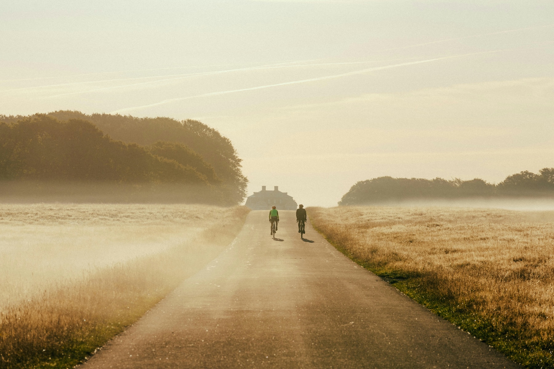 Two cyclists ride a misty dawn road towards the Hermitage Palace in Jægersborg Dyrehave, north of Copenhagen.