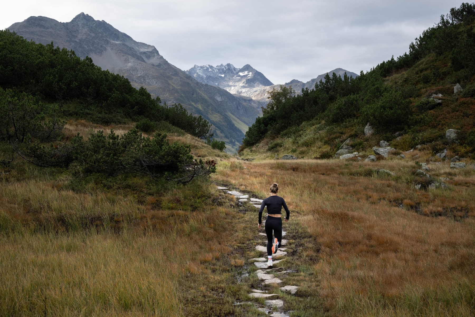 Runner on a stone path through golden alpine meadows below the peaks of the Verwalltal during the ULLR's Alpine Training Experience 1300, St. Anton | The Aficionados