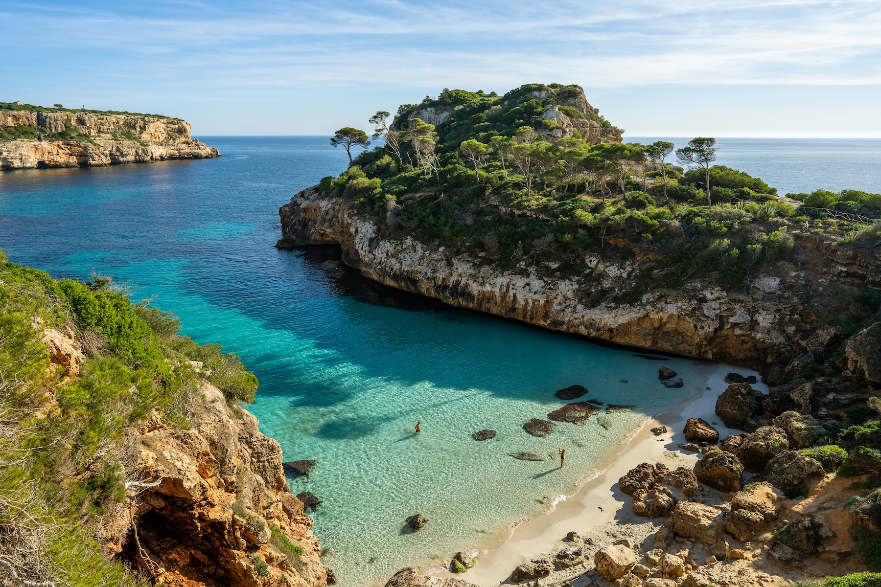 A turquoise cove on Mallorca's south-east coast near Santanyí, with pines on the limestone headland and two swimmers in the shallows | The Aficionados