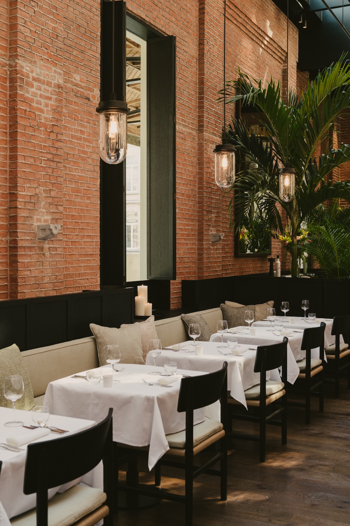 Red brick dining room with white-clothed tables, black chairs and a palm in daylight at August, Antwerp | The Aficionados