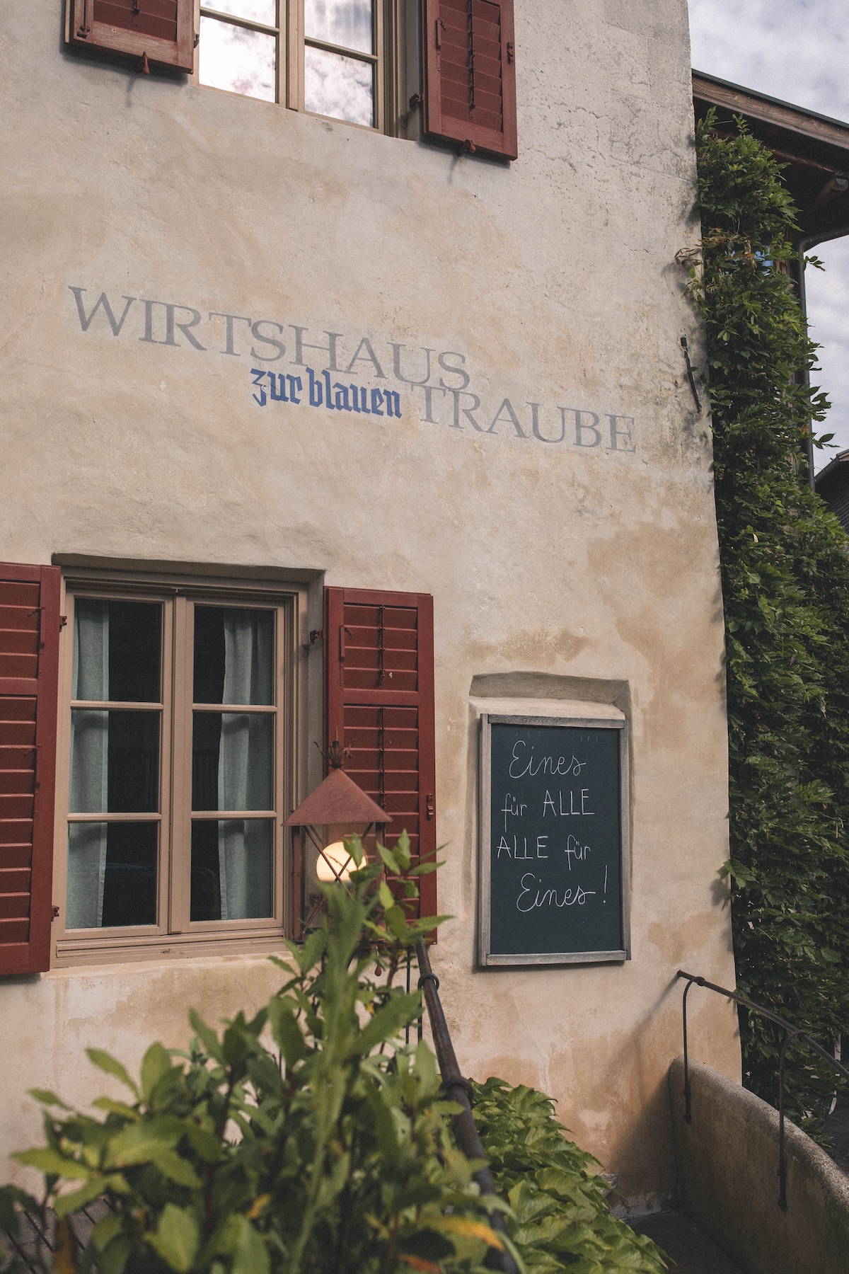 Painted ochre facade of Wirtshaus zur Blauen Traube in Algund, South Tyrol, with rust-red shutters, climbing ivy and the chalkboard credo Eines für ALLE, ALLE für Eines | TheAficionados.com