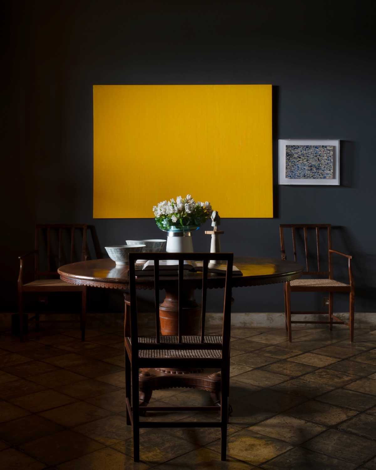 Vivid yellow monochrome painting on a charcoal-hued wall above a round antique dining table with carved wooden chairs and ceramic bowls, from Peter Harris's contemporary art collection at Doornberg, Galle, Sri Lanka | The Aficionados.