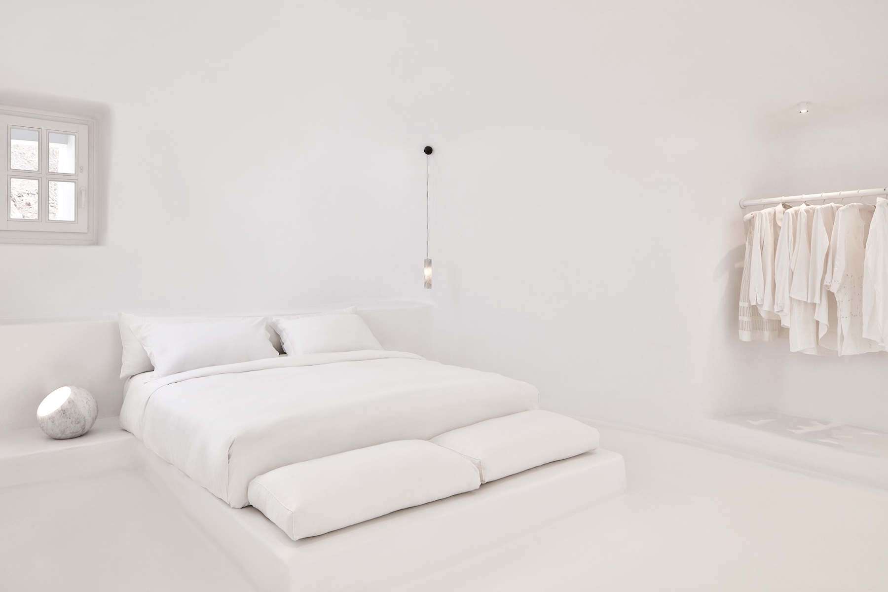 A white-on-white Cycladic bedroom at Aenaon Villas, Santorini, with a moulded plaster bed platform, a spherical marble lamp and a rail of pale linen.