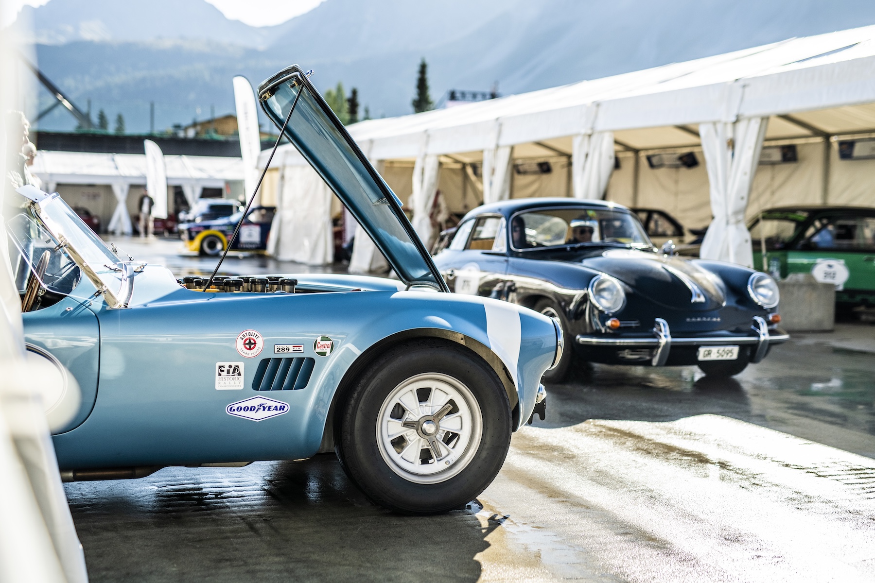 Light-blue Shelby Cobra 289 with bonnet raised beside a black Porsche 356 in the Arosa ClassicCar paddock | The Aficionados