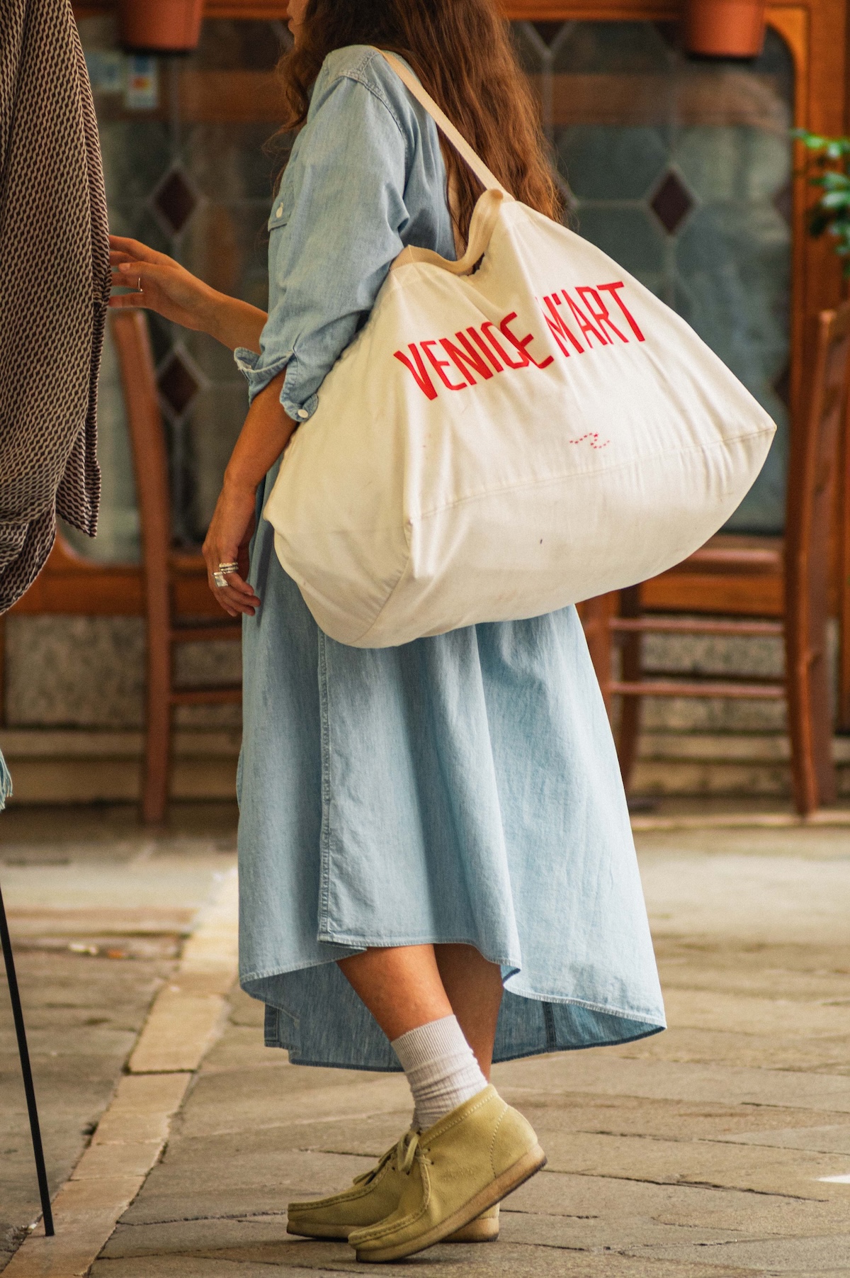 A woman in a denim shirt-dress carries an oversized cream canvas Venice M'Art tote with bold red lettering over her shoulder.