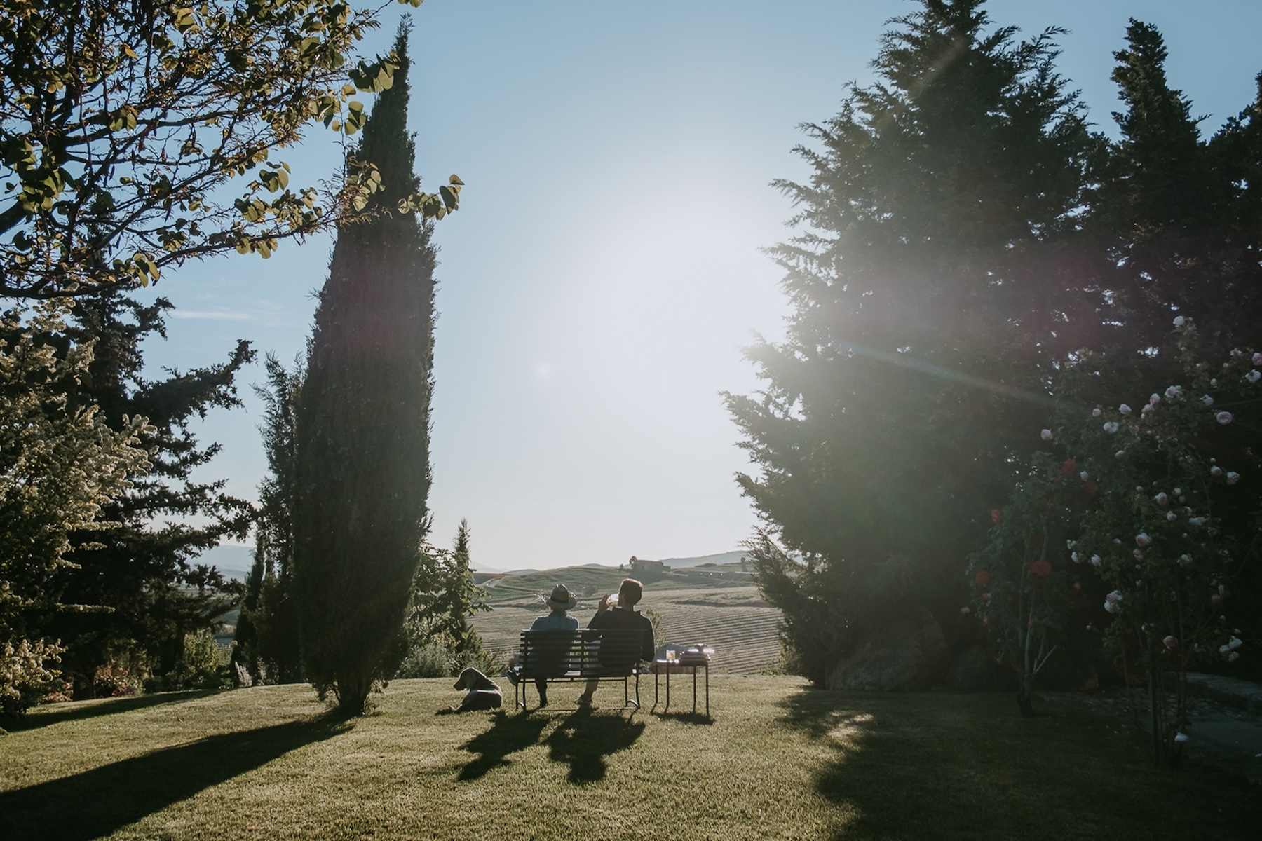 Golden-hour view on the lawn at Susafa, guests on a garden bench with the family dog and the rolling Sicilian countryside stretching toward the Madonie foothills, country boutique hotel above Polizzi Generosa | The Aficionados.