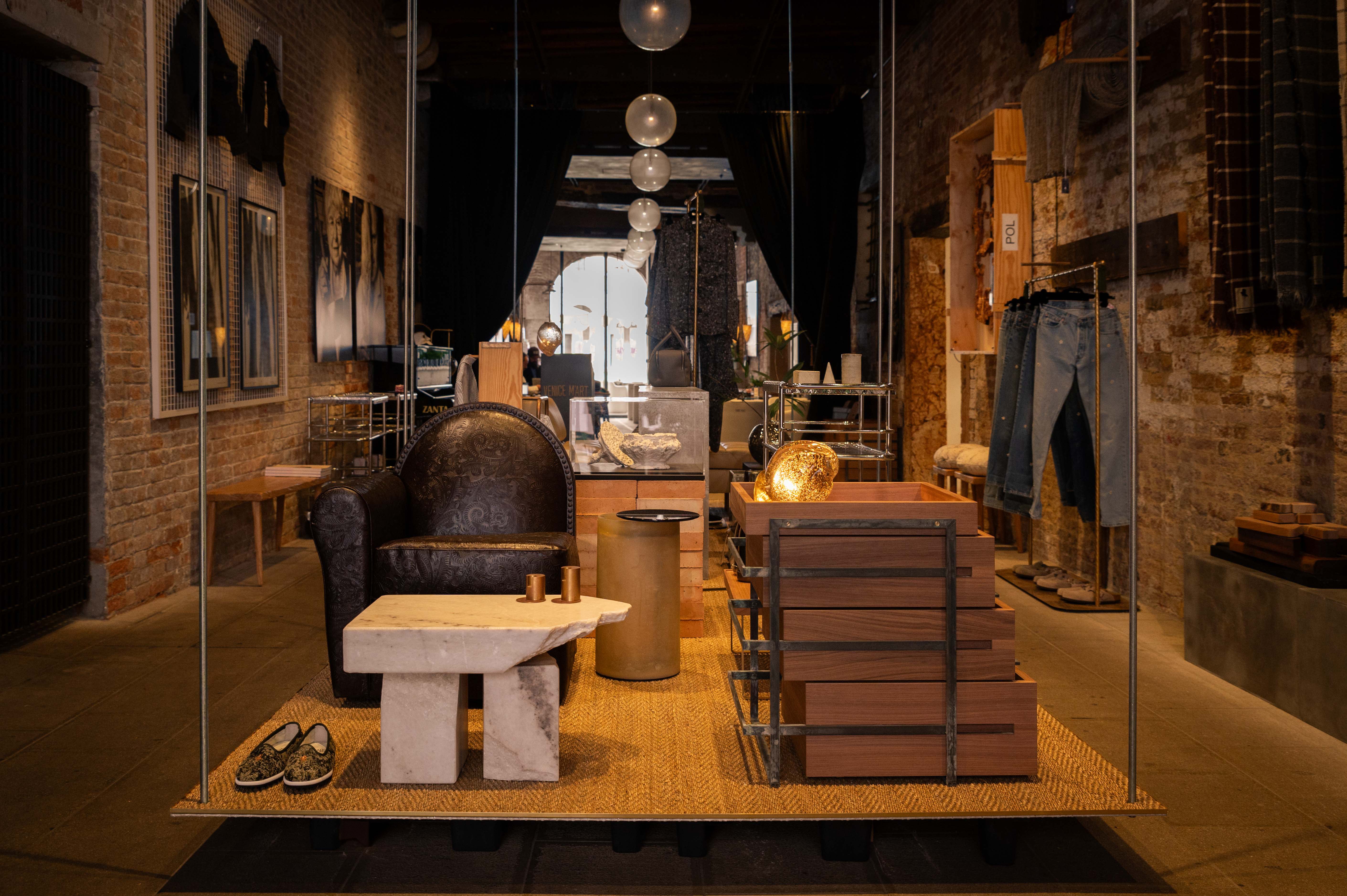 Interior of Venice M'Art inside The Venice Venice Hotel: exposed brick walls, a paisley leather armchair, a marble coffee table and stacked walnut crate-tables under glass pendant lights, with denim and artwork on display.