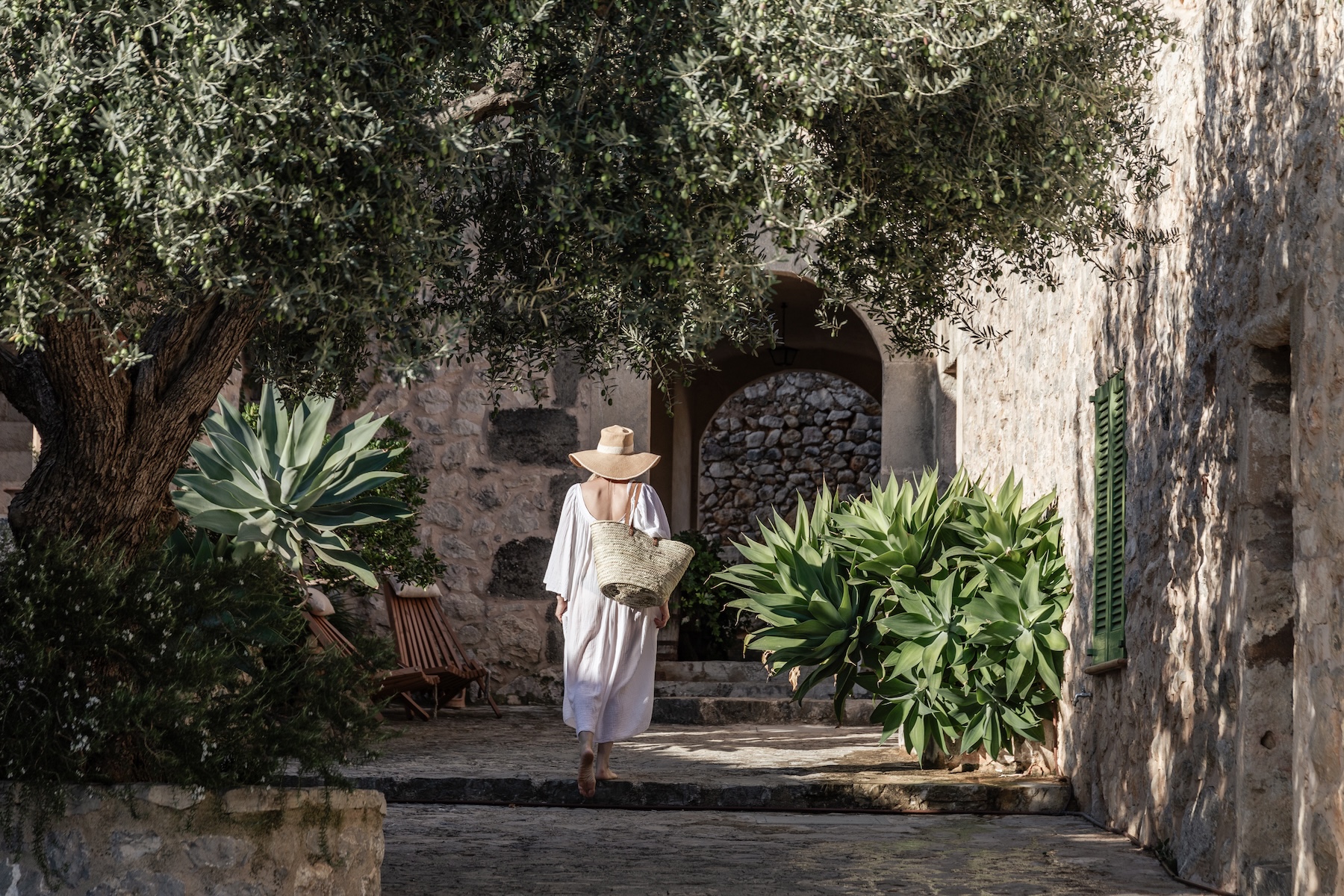 A guest in white crosses the olive-shaded stone courtyard at Ca'n Beneït, Mallorca, framed by agaves and green shutters.