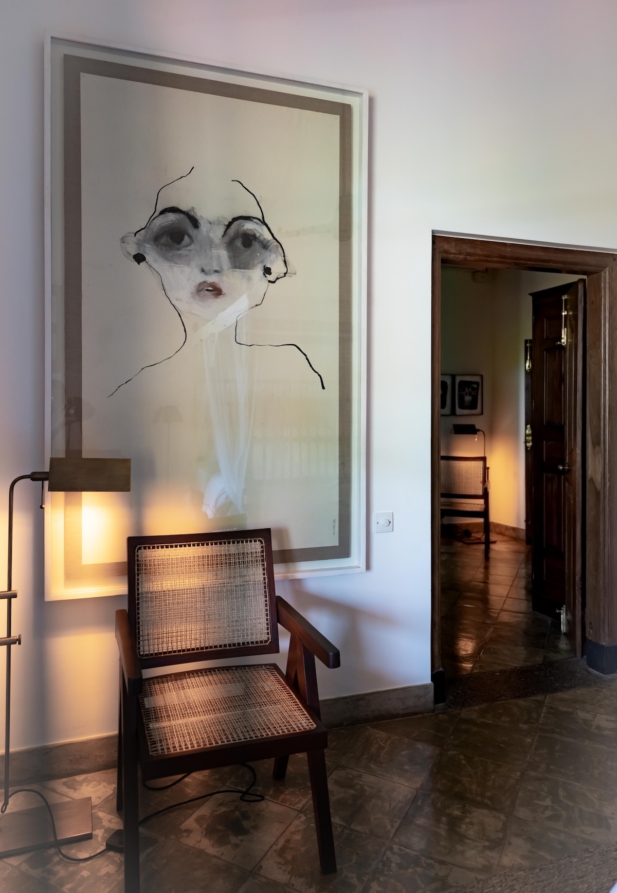 Large framed expressive face drawing in soft greys and ink, hung above a Pierre Jeanneret-style cane armchair and brass floor lamp in a hallway at Doornberg, Peter Harris's art-filled villa, Galle, Sri Lanka | The Aficionados.