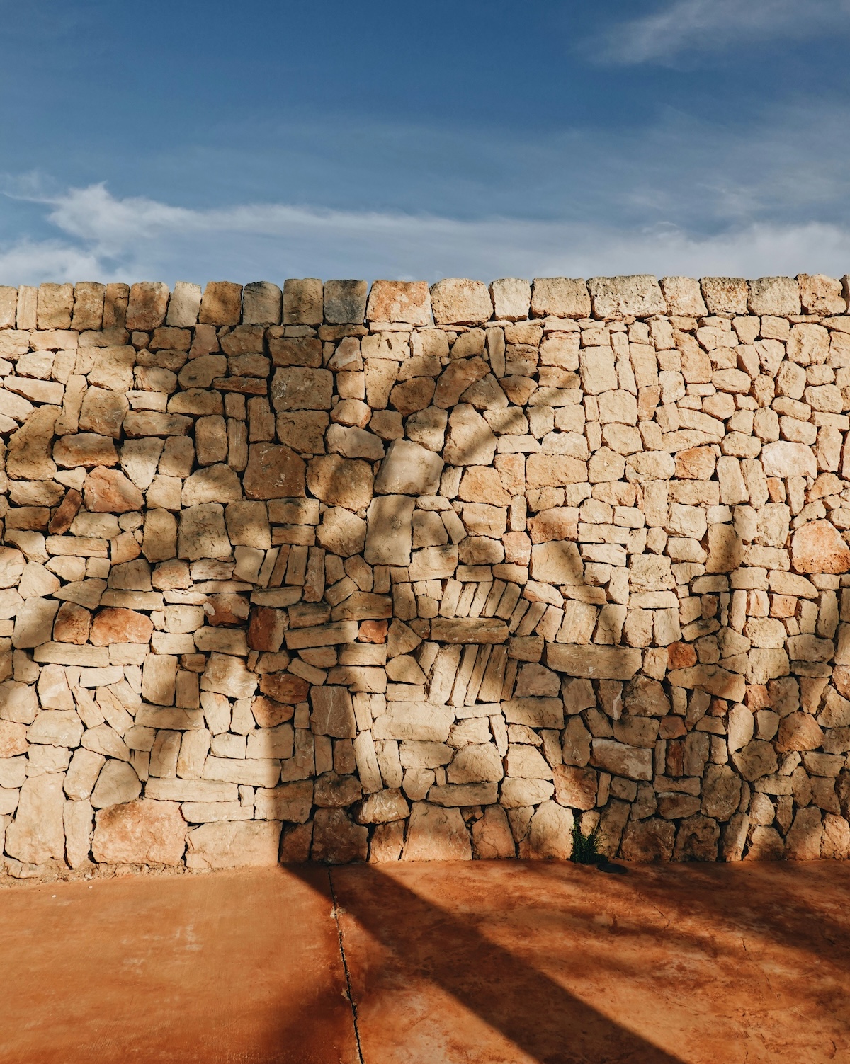 A Mallorcan dry stone wall of honey limestone above a terracotta floor, with the shadow of a palm falling across it | The Aficionados