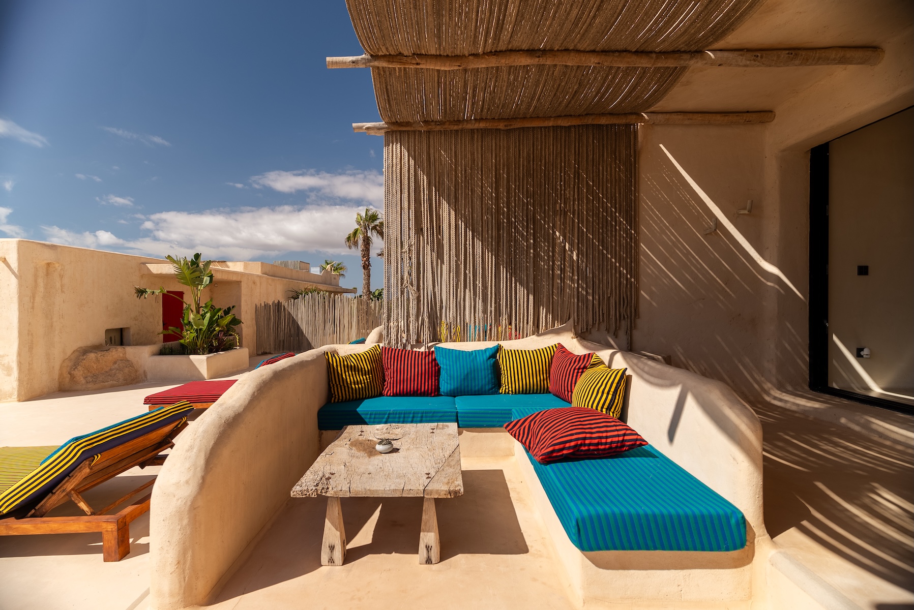 A curving whitewashed terrace at Hannah, Formentera, with a rope shade and a built-in banquette dressed in bold blue, red and yellow striped cushions.