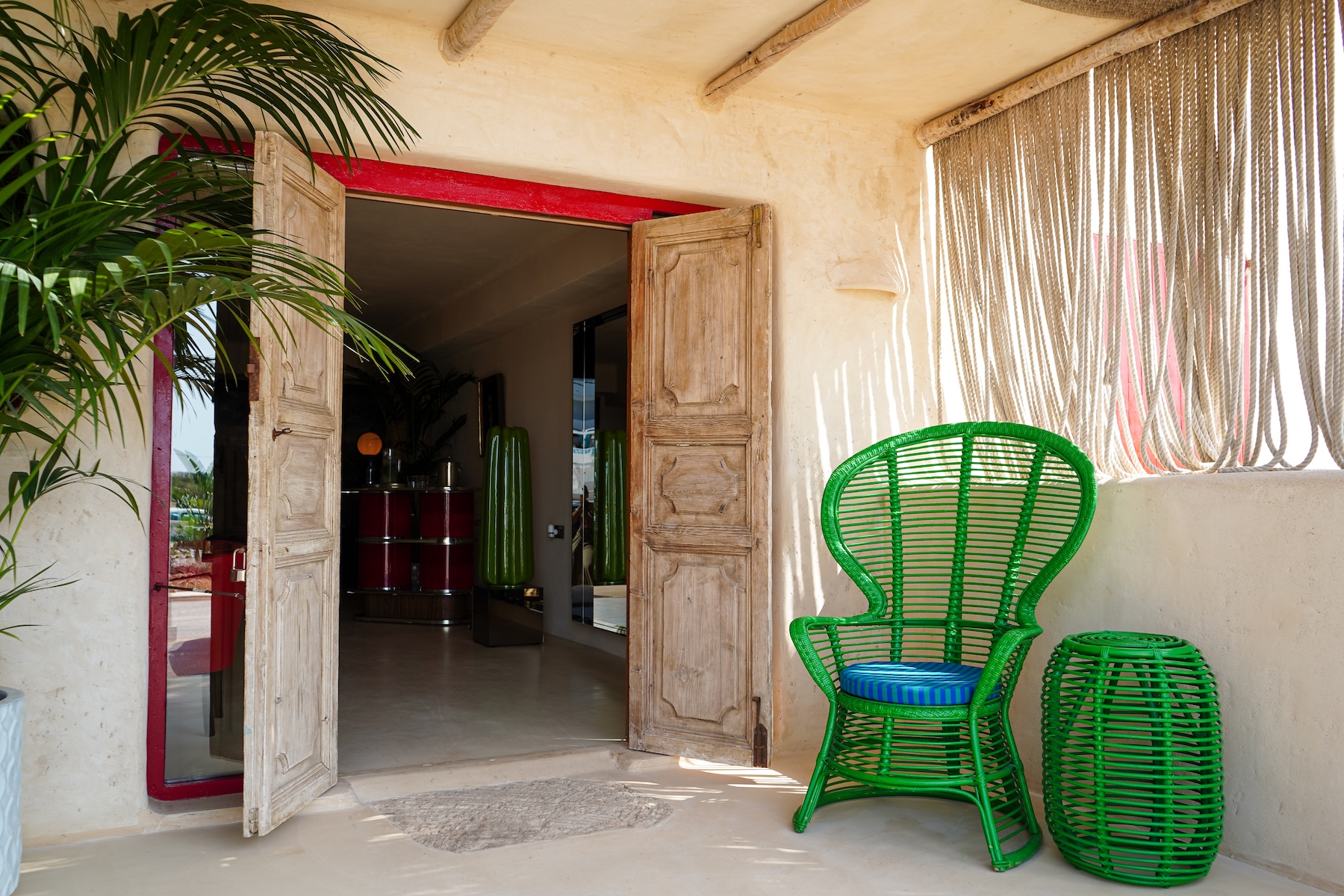 Guest room entrance at Hannah Formentera with a red-framed door, weathered wooden shutters and a green rattan peacock chair | The Aficionados