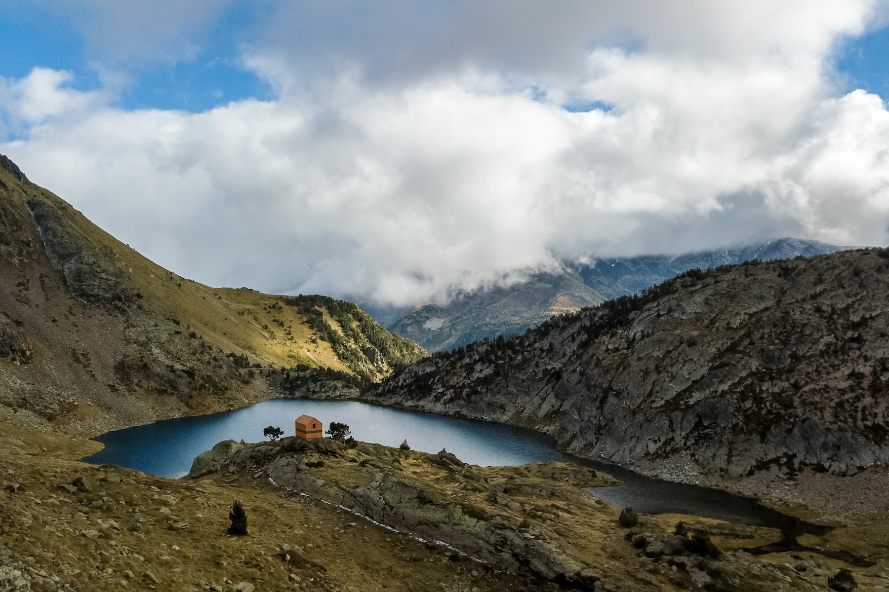 Timber refuge hut on a rock spur beside a high mountain lake, cloud sitting on the ridges beyond, Andorra
