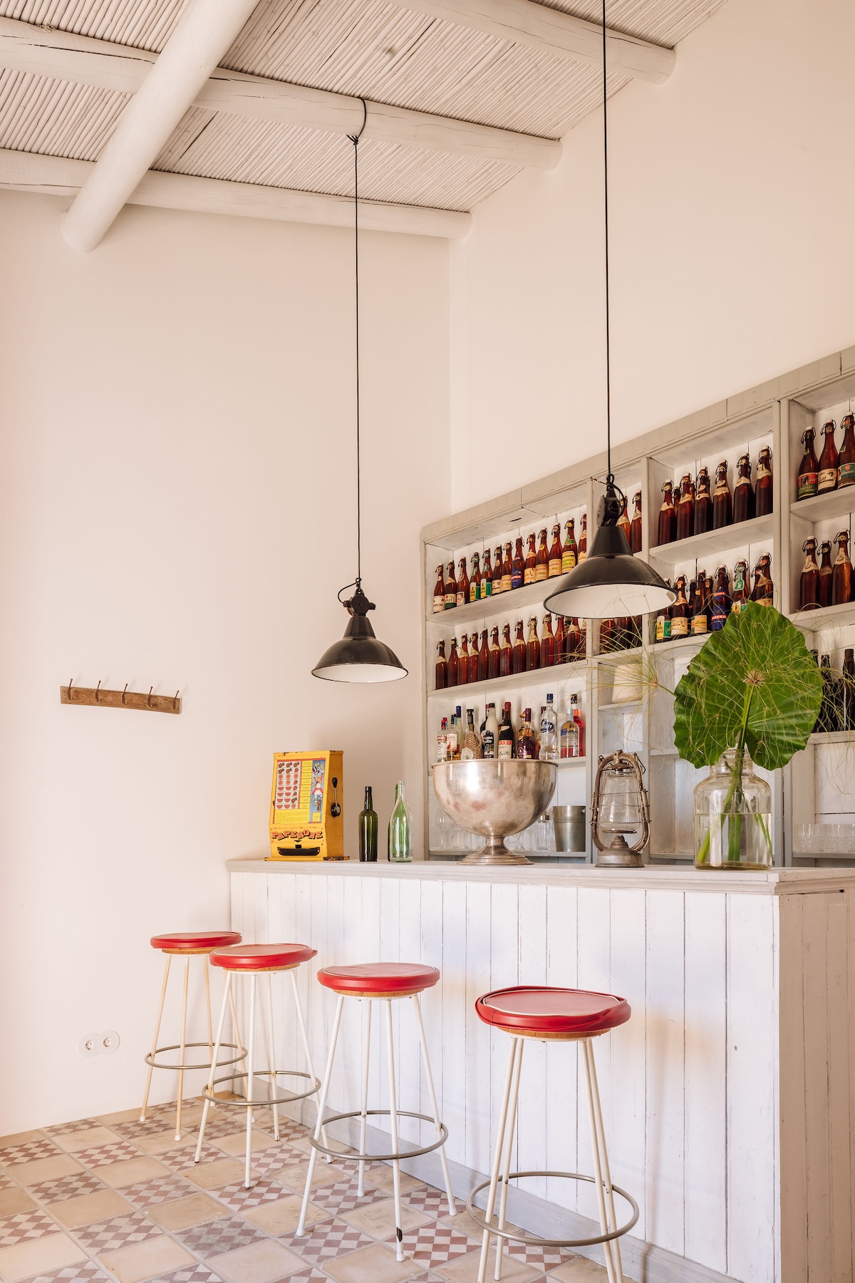 The bar at Hospedaria in Tavira, with a whitewashed plank counter, red-topped stools, industrial pendant lamps and shelves stacked with bottles above a patterned tiled floor.