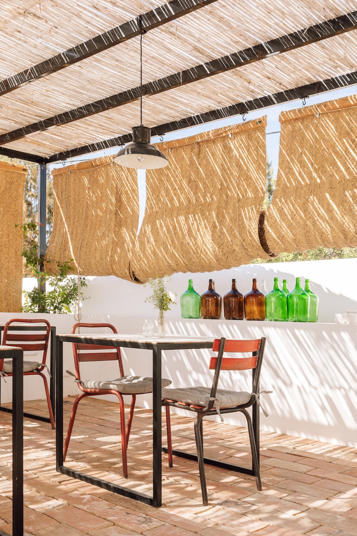 Sunlit terrace corner at Hospedaria in Tavira with rust-red metal chairs, a wooden table and esparto roller blinds, lined with green and amber glass demijohns.