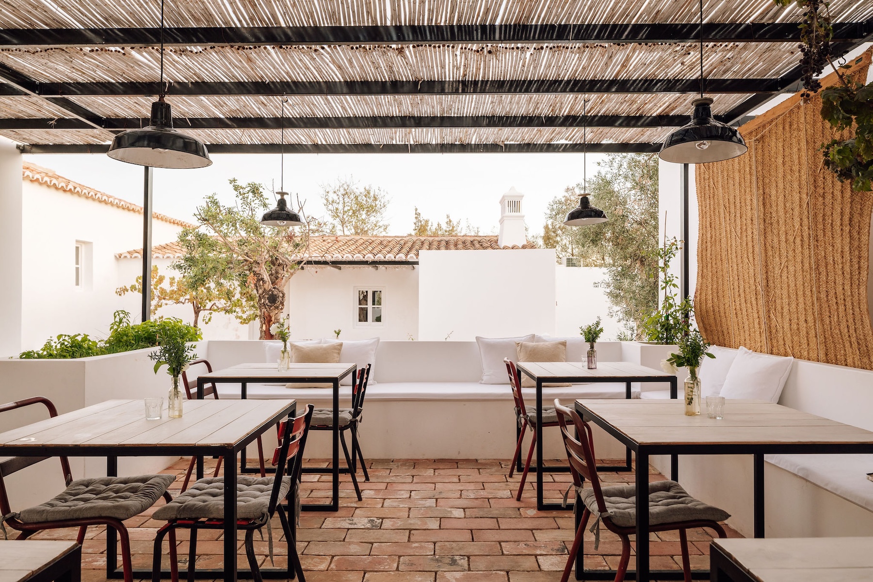 Outdoor dining terrace at Hospedaria in Tavira, with an esparto reed pergola, black enamel pendant lamps, a white plaster banquette and wooden tables on a terracotta tiled floor, set against whitewashed Algarve buildings and an olive tree.