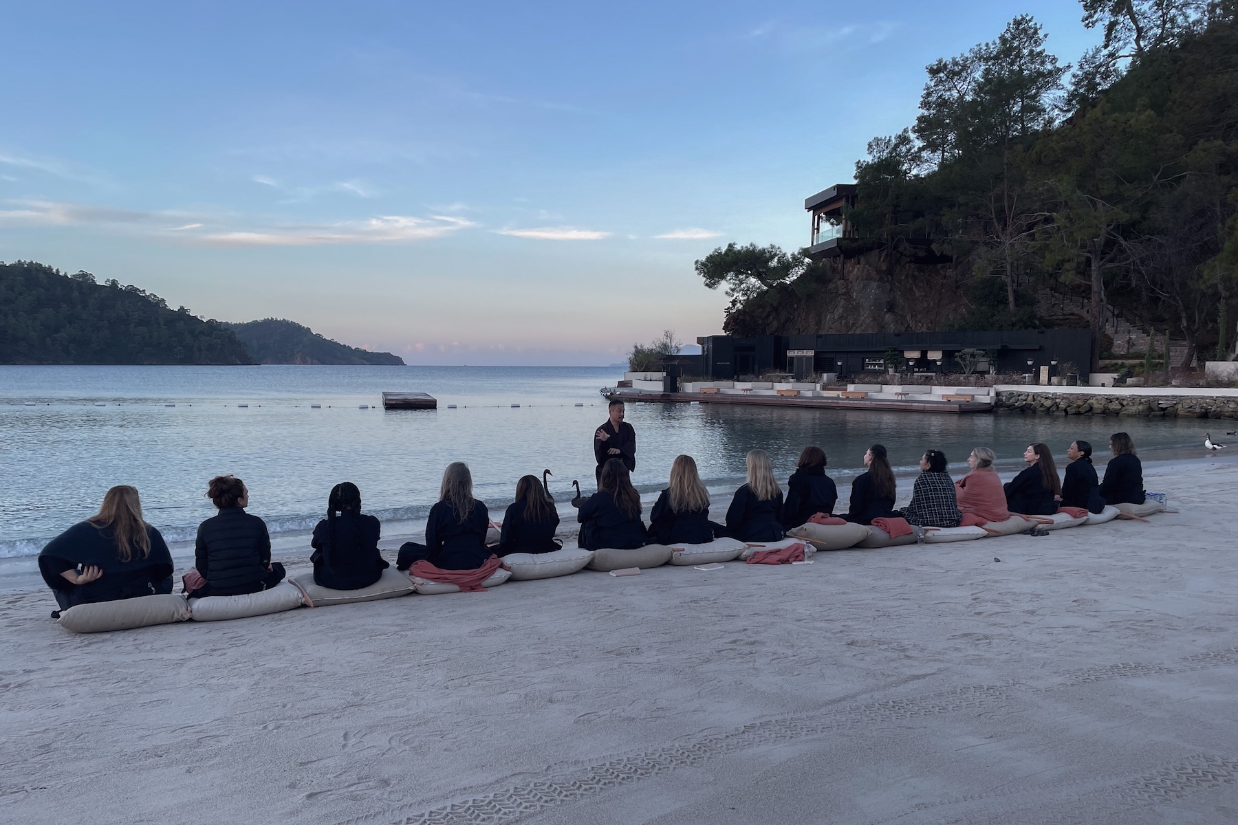 Participants seated on cushions for a sunrise meditation on the beach at AHAMA, facing the calm bay and forested hills of the Turkish Riviera.