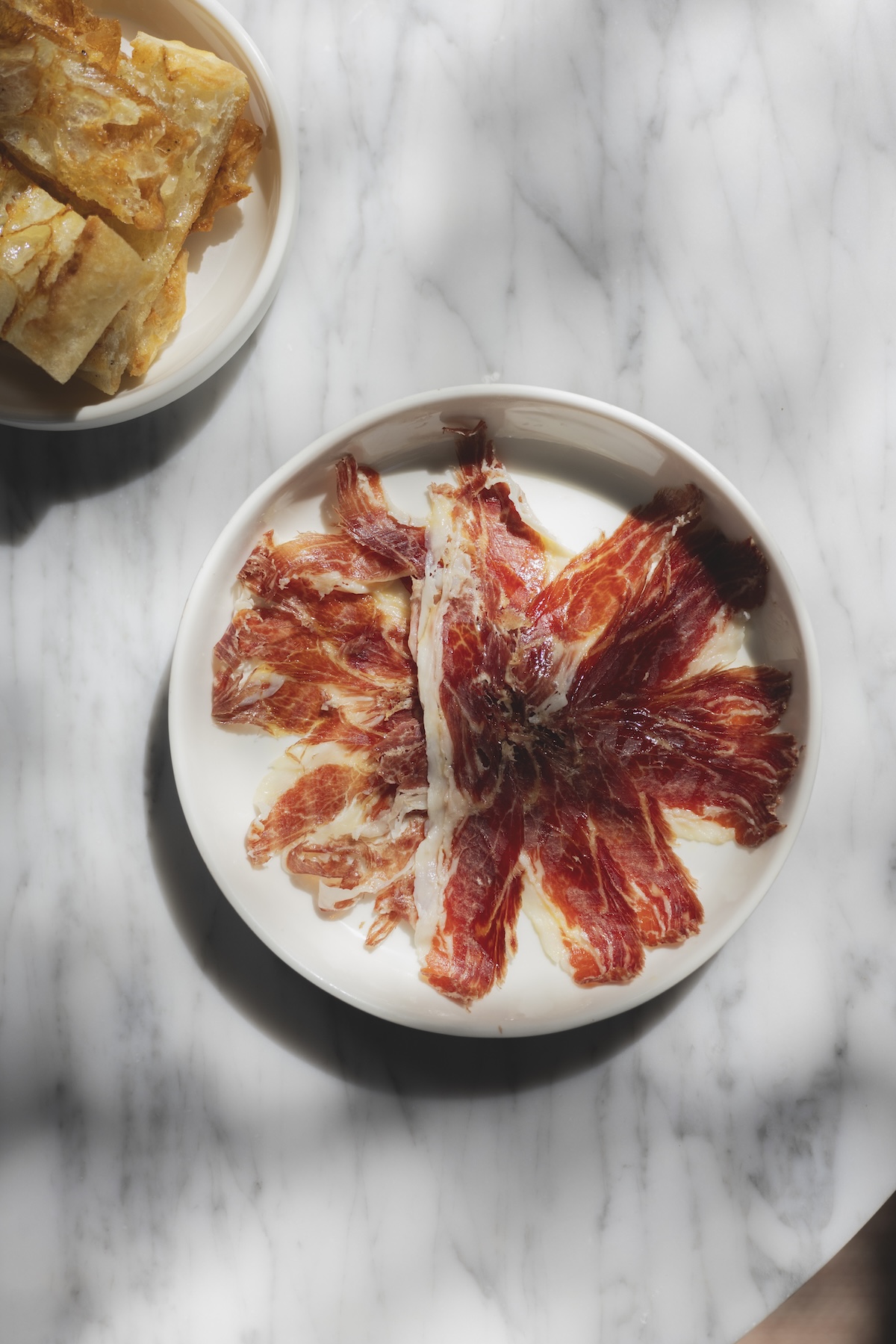 Plate of thinly sliced Iberian ham served with fried bread at August, Antwerp | The AficionadosRed brick dining room with white-clothed tables, black chairs and a palm in daylight at August, Antwerp | The Aficionados