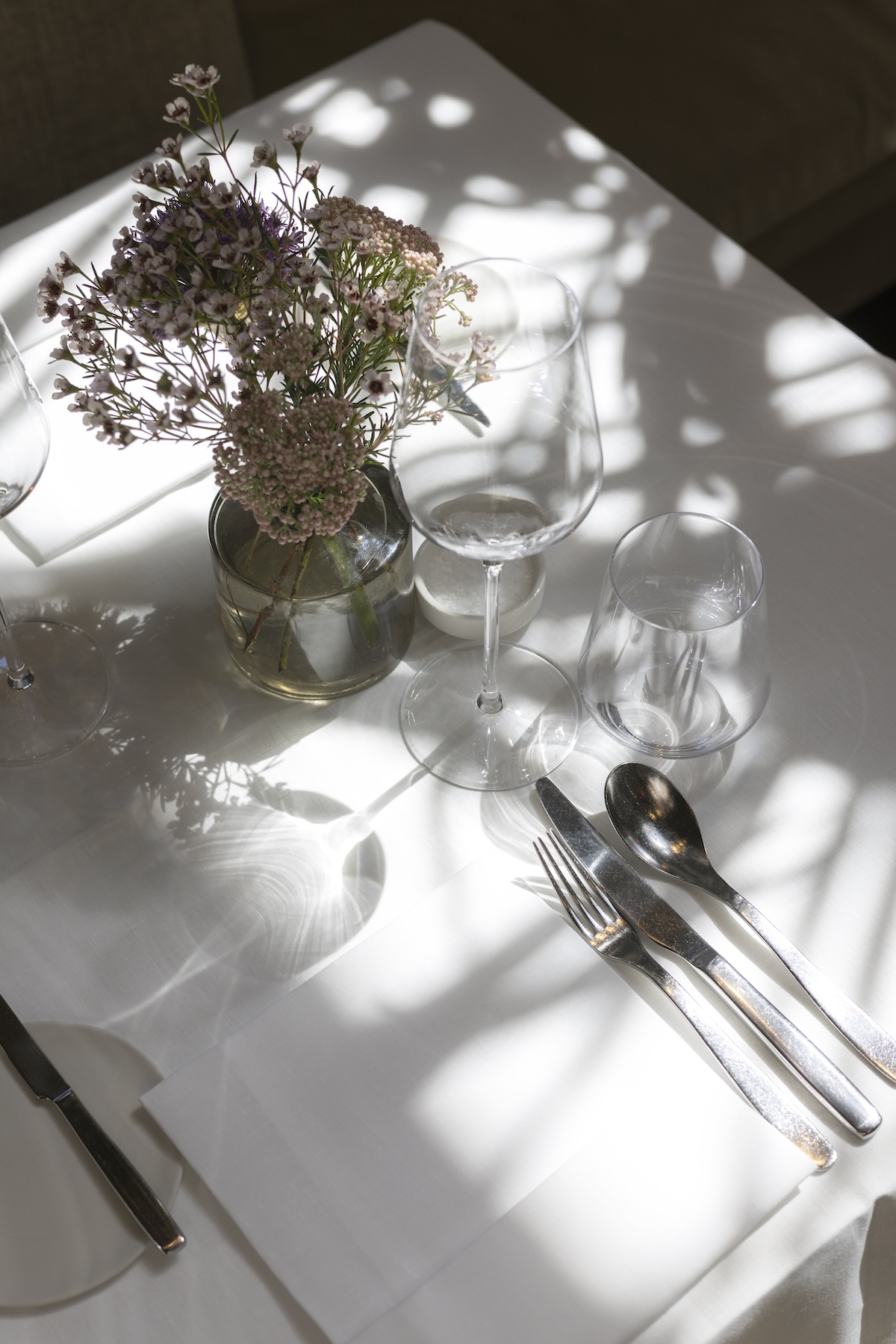 Sunlit table setting with a vase of waxflower, wine glasses and cutlery on white linen at August, Antwerp | The Aficionados