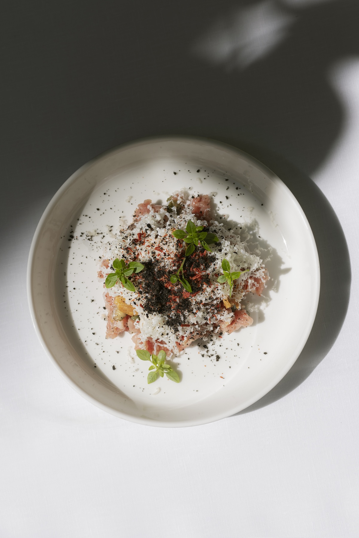 Tartare finished with grated cheese, herbs and dark crumb on a white plate at August, Antwerp | The Aficionados