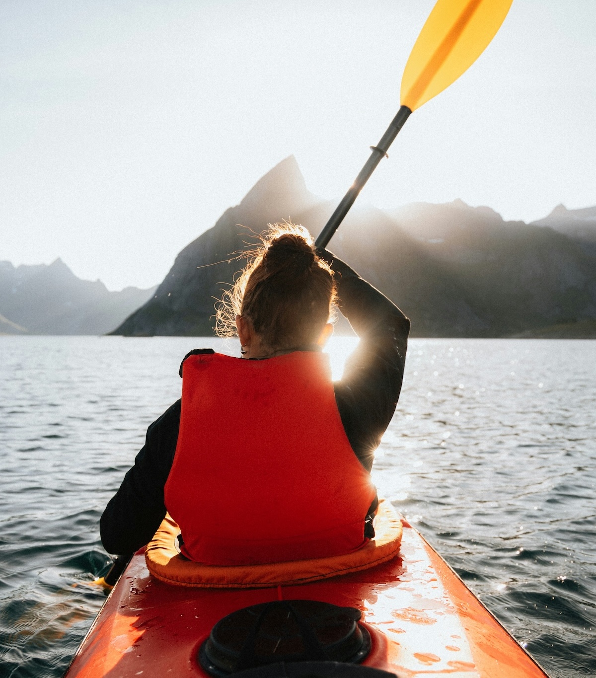 Sea-kayaking on calm Arctic water at Nusfjord, Lofoten, paddle raised against a low backlit sun and mountains | The Aficionados