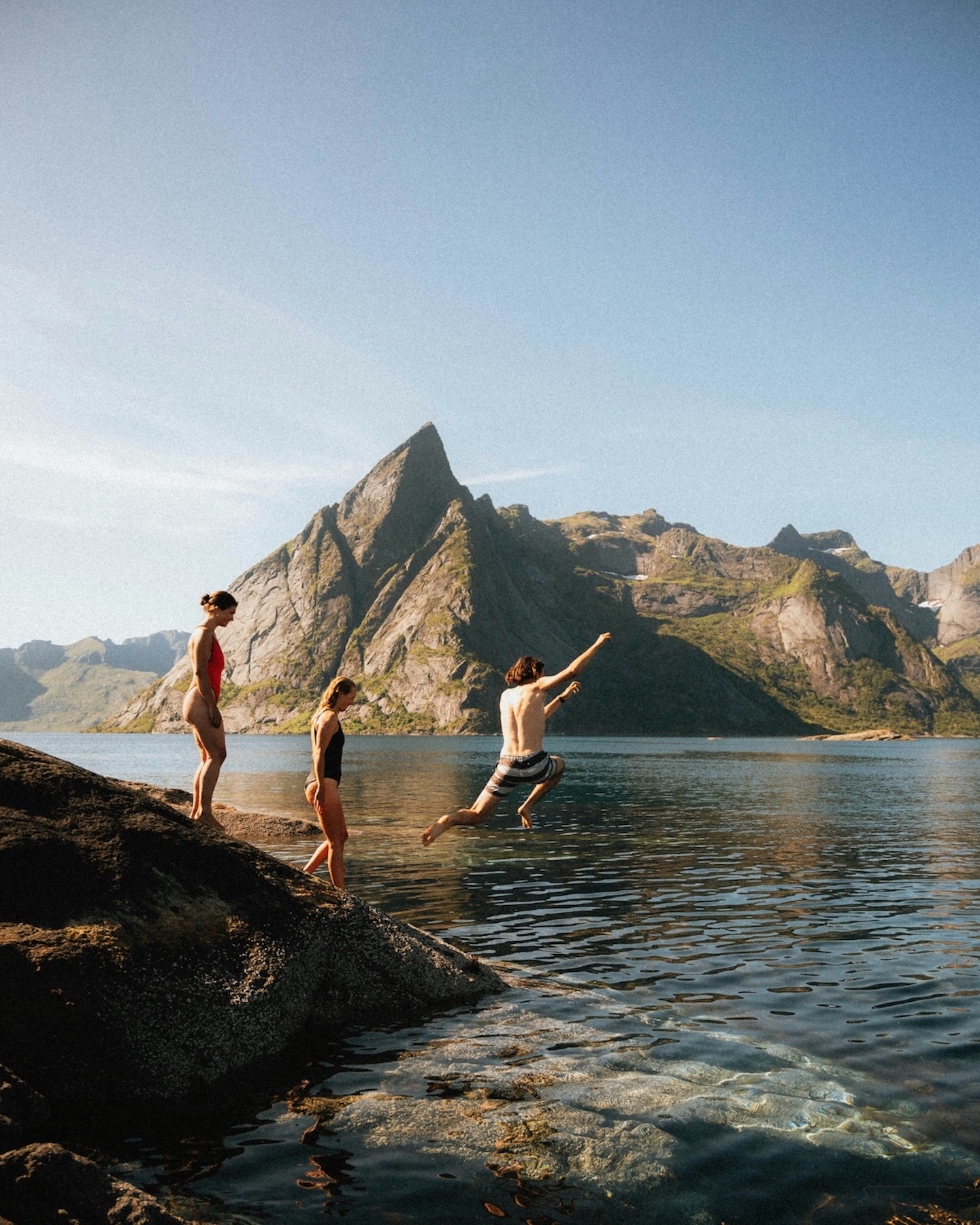 A surfer in a wetsuit holds a longboard upright on the shore at Flakstad Beach, Lofoten, mountains rising behind | The Aficionados