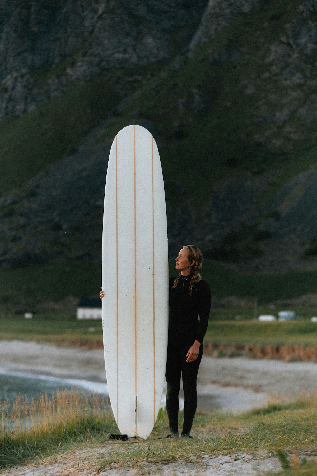 A surfer in a wetsuit holds a longboard upright on the shore at Flakstad Beach, Lofoten, mountains rising behind | The Aficionados