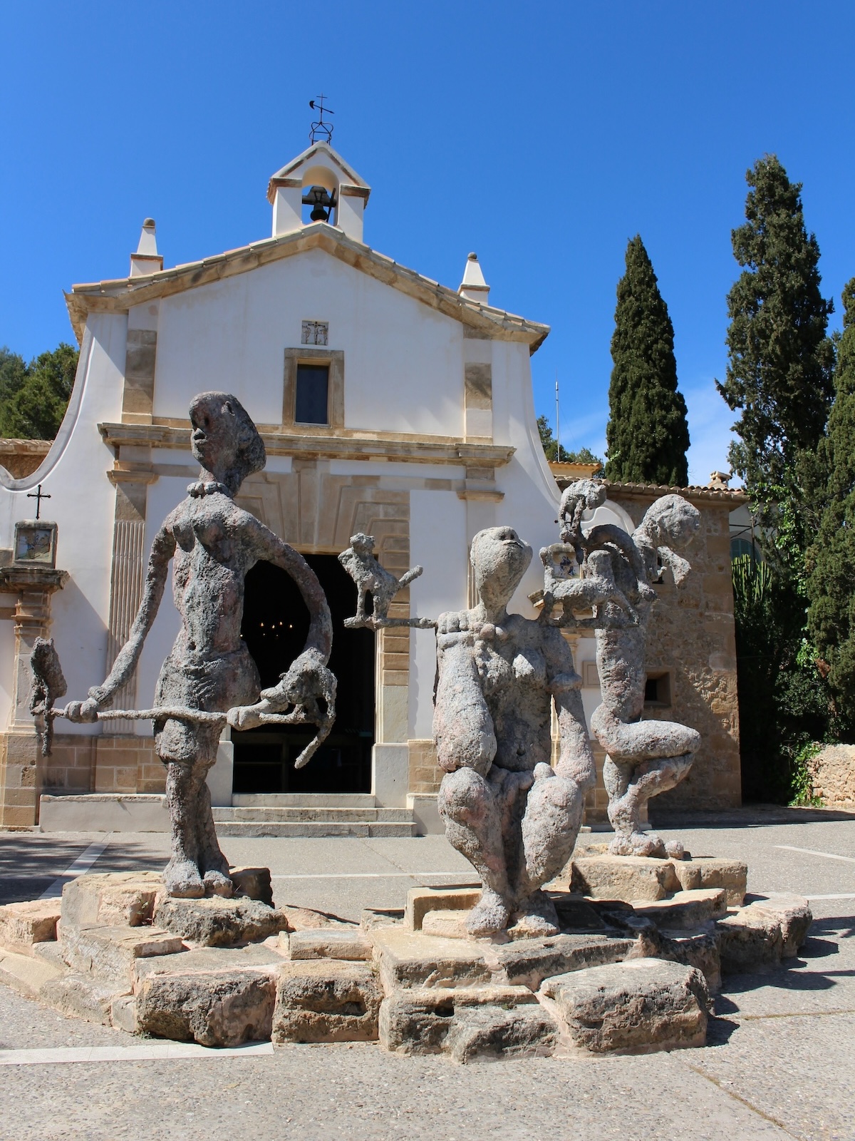 Rough-textured bronze figures grouped on stone slabs before a whitewashed Mallorcan chapel and cypresses | The Aficionados