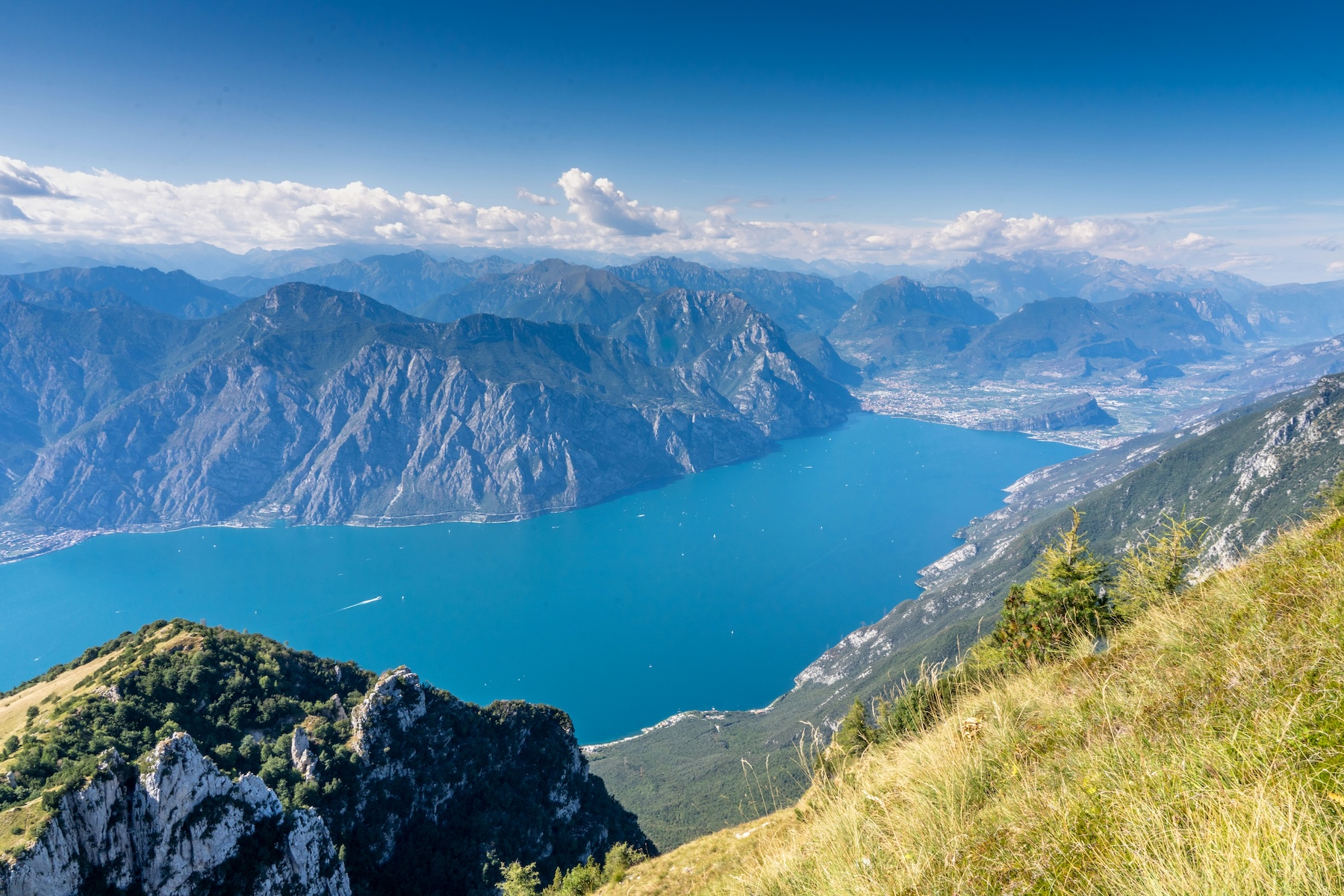 The northern reach of Lake Garda seen from a high ridge, turquoise water hemmed by limestone cliffs with Riva del Garda and Torbole at the lake head and the Brenta peaks beyond | The Aficionados