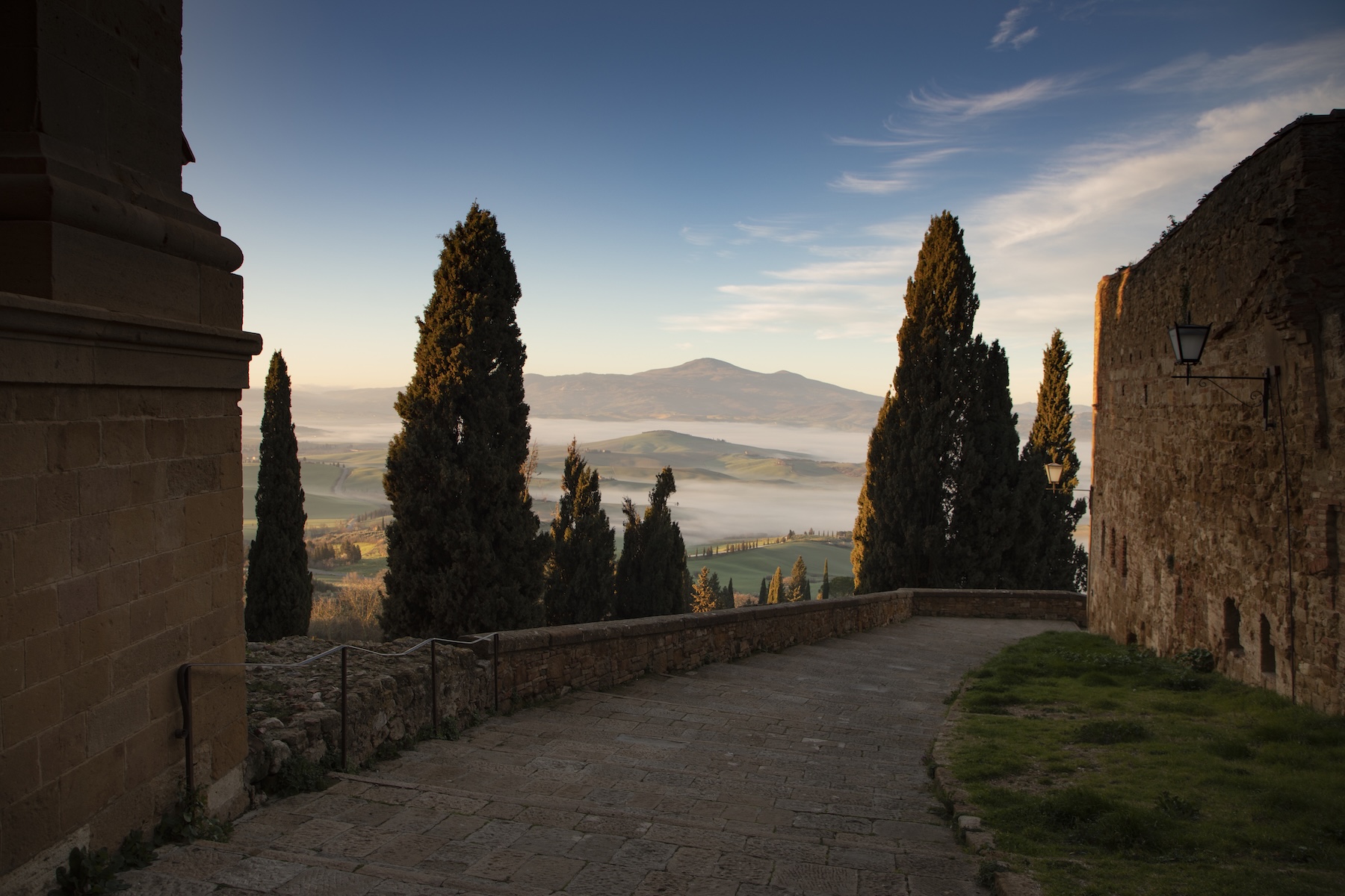 PIENZA | La Bandita Townhouse Hotel | Cypress trees framing a misty Val d'Orcia valley at dawn from a Tuscan hilltown lane