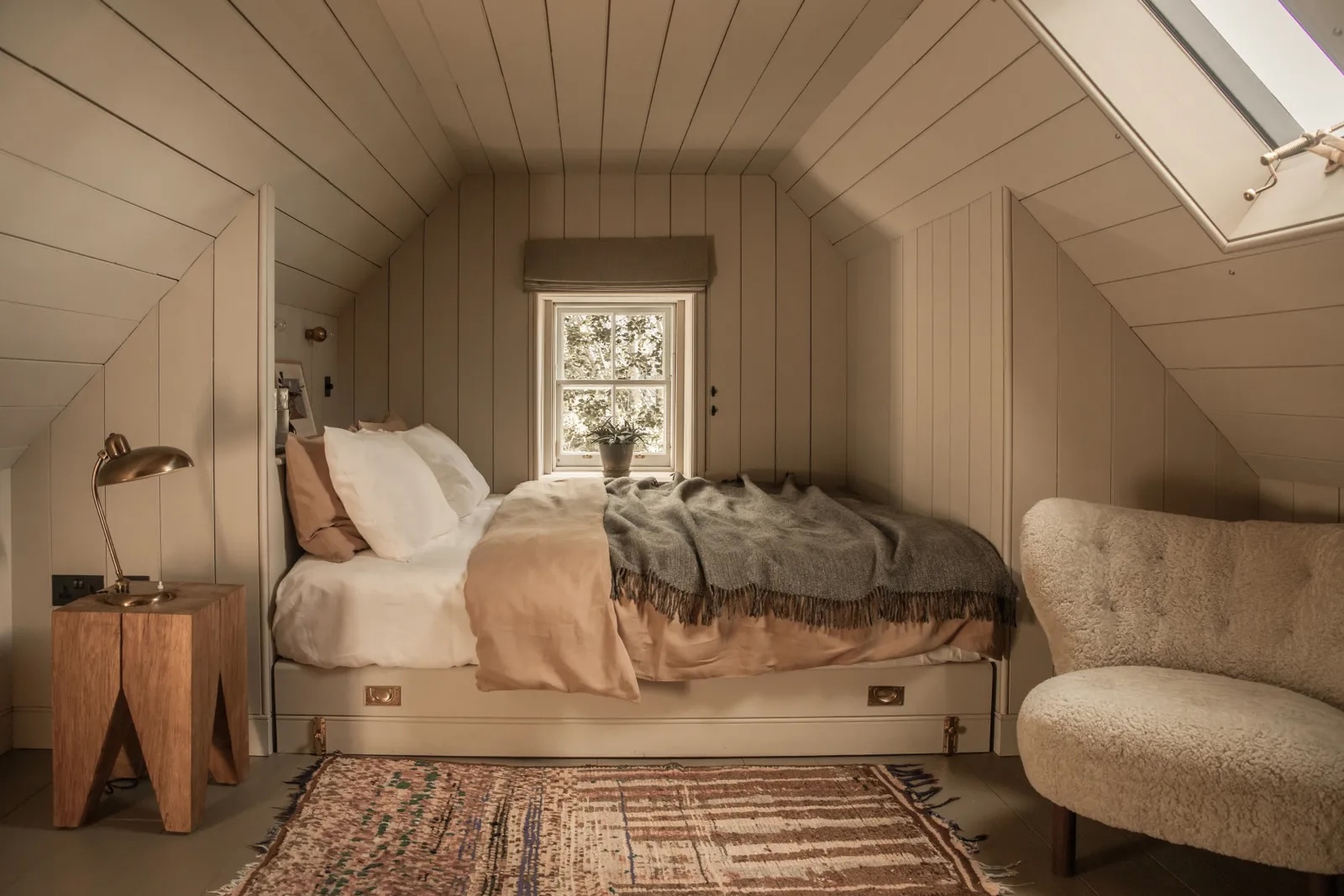 Attic bedroom at Lundies House, Tongue, with pale timber panelling, a skylight, sheepskin chair, brass lamp and Scandi-Scot styling in soft neutral tones.