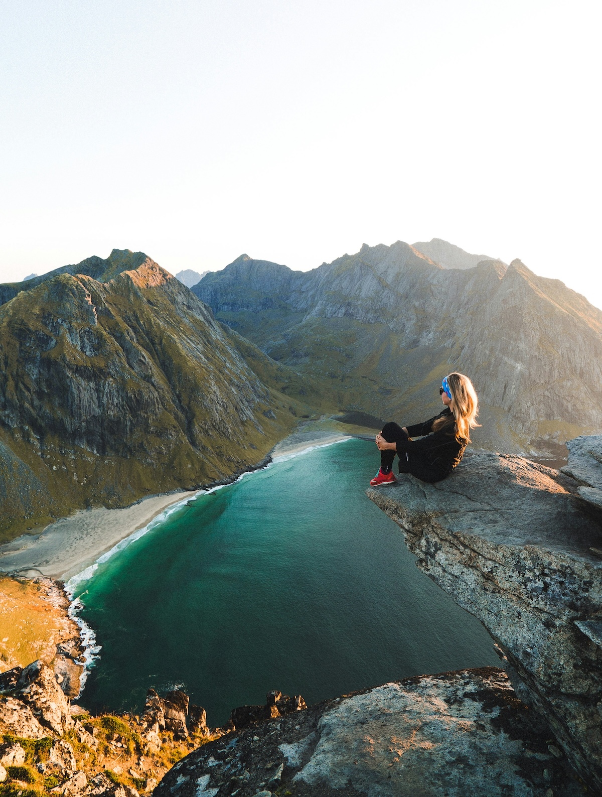 A hiker rests on a clifftop above a turquoise Lofoten beach and ridgelines, on a guided mountain hike near Nusfjord | The Aficionados