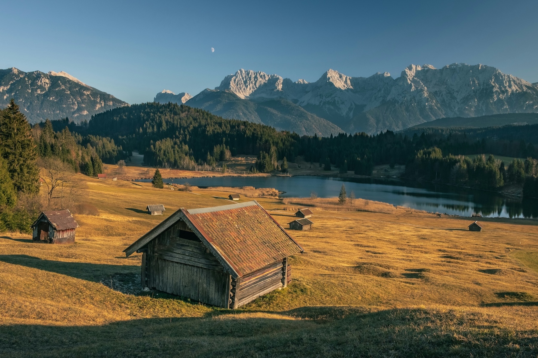 Geroldsee, near Krün | Wooden hay huts in a golden autumn meadow by a calm lake below the Bavarian Alps at dusk, a moon rising | The Aficionados.
