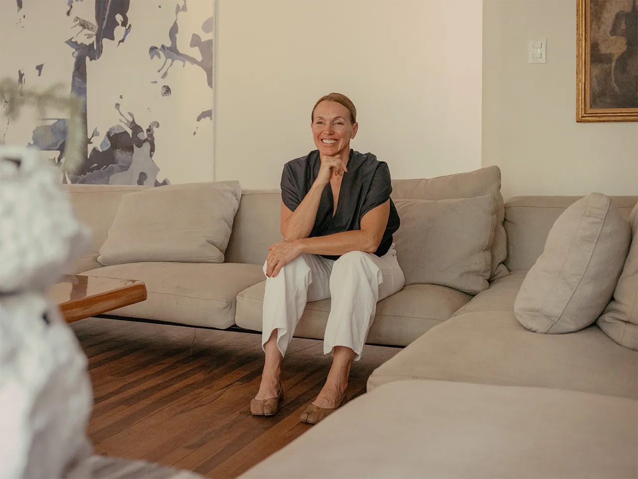 Mexican architect Tatiana Bilbao, founder of Tatiana Bilbao Estudio, seated on a pale linen sofa in a black top and white trousers, hand to her chin, smiling, with abstract artwork behind her.