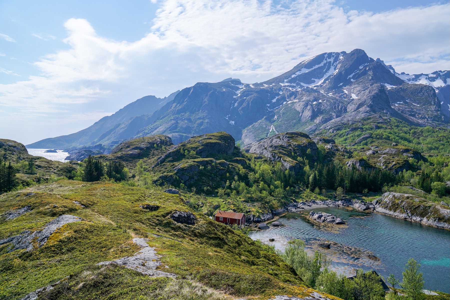 A lone red cabin beside a still inlet below snow-streaked peaks in summer, Lofoten, near Nusfjord Resort & Village | The Aficionados