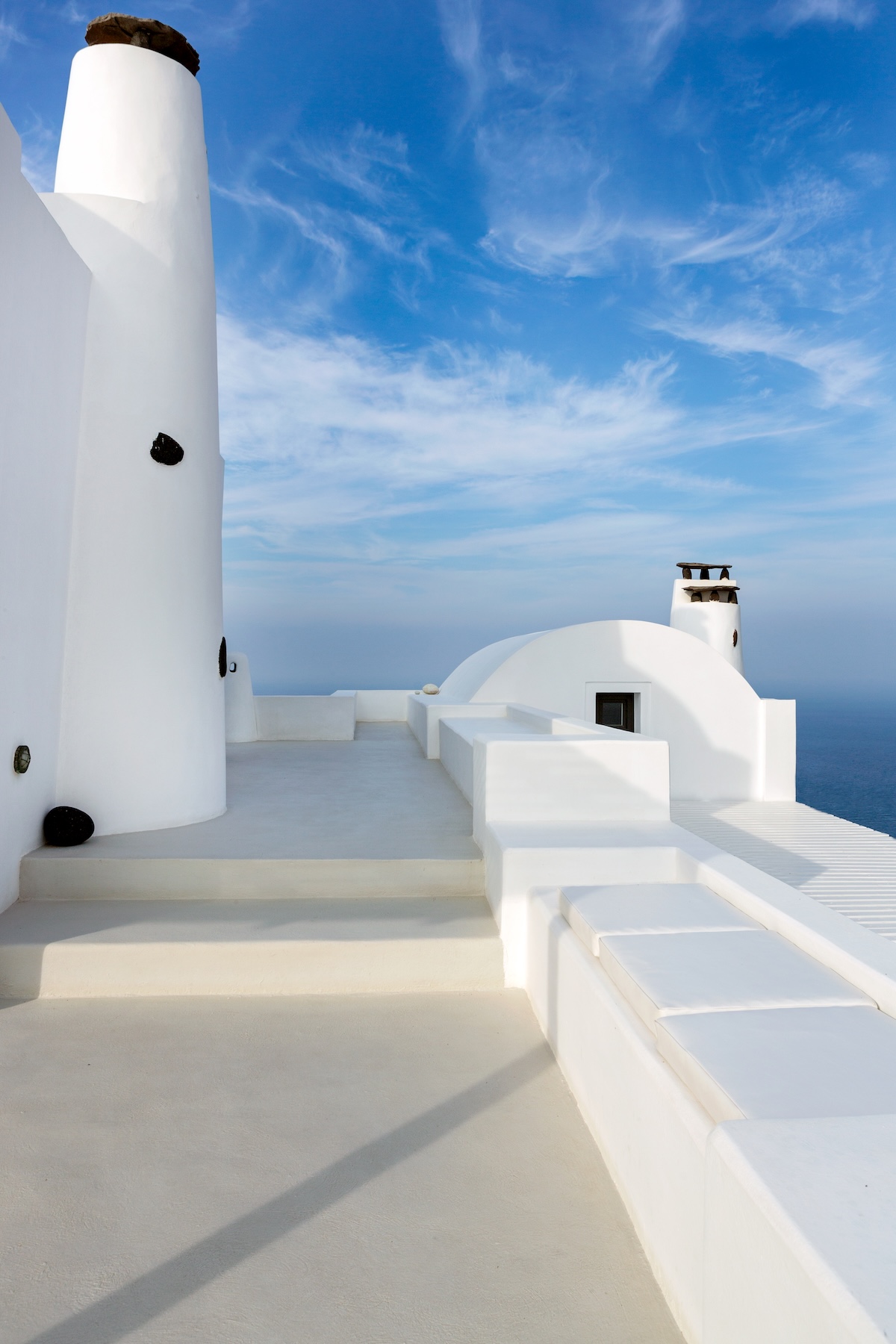 Whitewashed Cycladic chimneys and a domed roof against blue sky and the Aegean at Aenaon Villas, boutique design villas on the edge of Santorini's caldera.