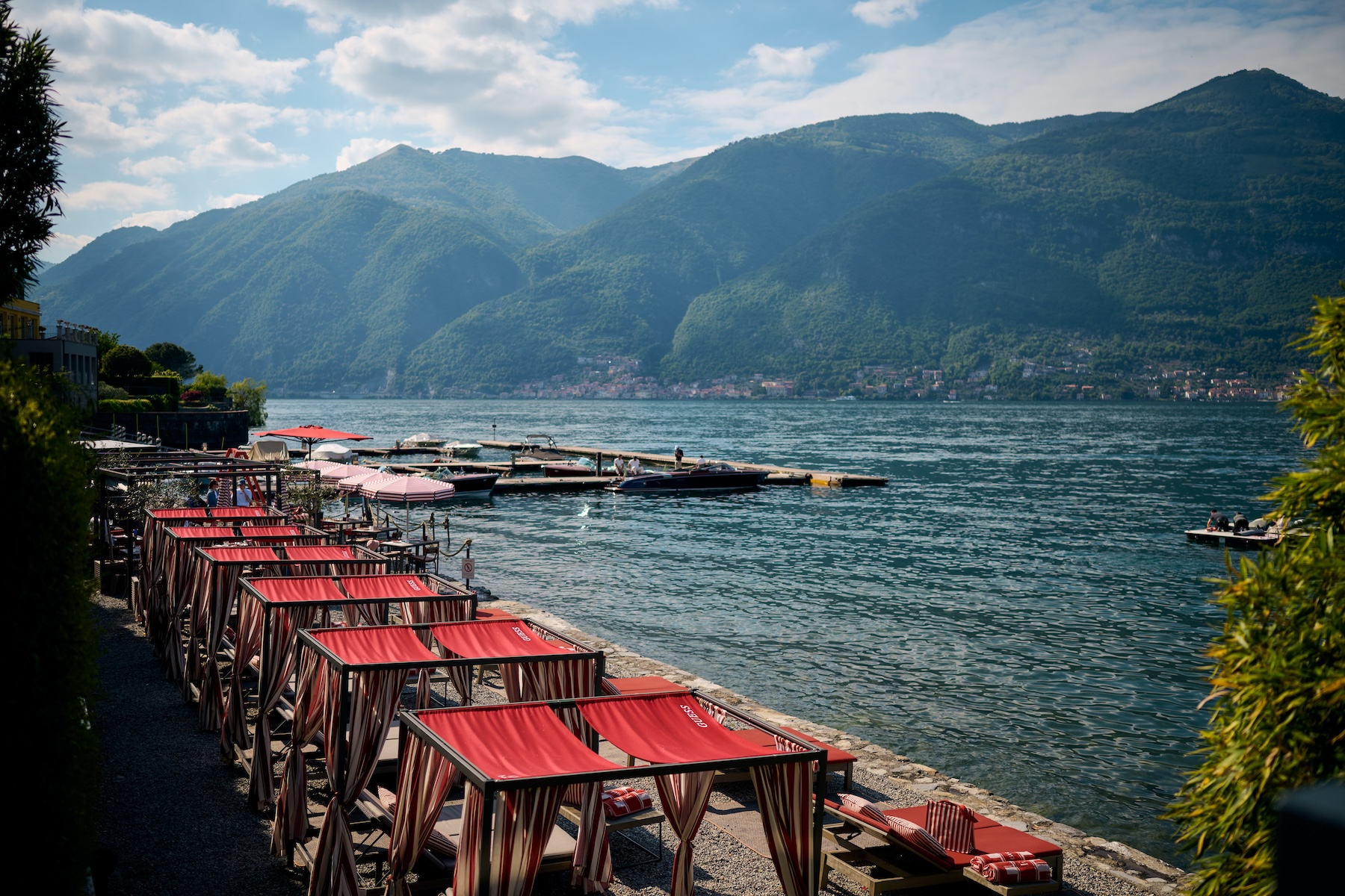 Filario Hotel lakeside lido with row of red GUESS cabanas, Lezzeno shoreline, private marina, Lake Como mountain views, Ciao Como summer 2026 collaboration | TheAficionados.com