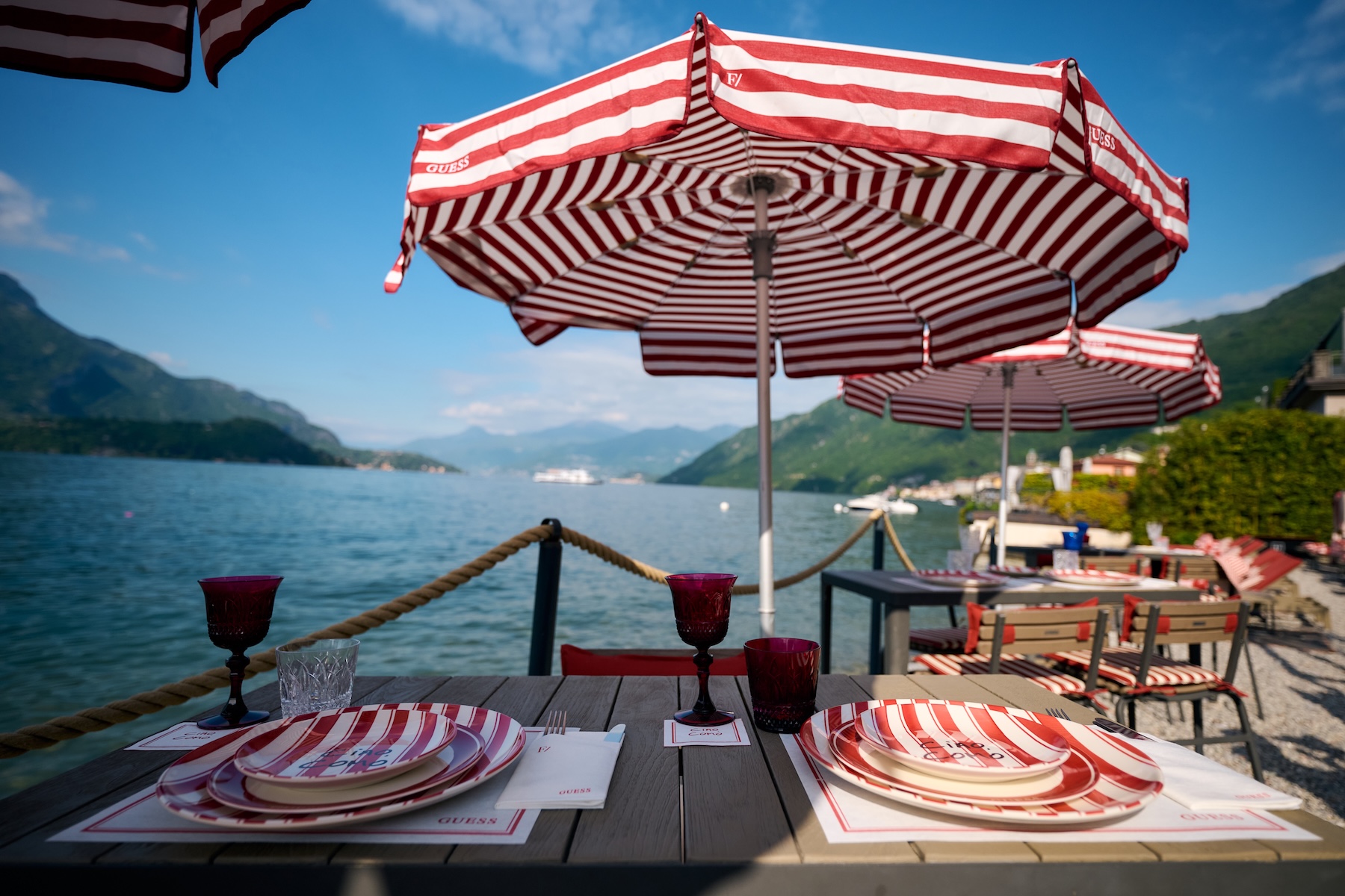 Filario lakefront restaurant terrace table setting, striped GUESS Ciao Como umbrellas, dinnerware, placemats and ruby wine glasses, Lake Como dining | TheAficionados.com