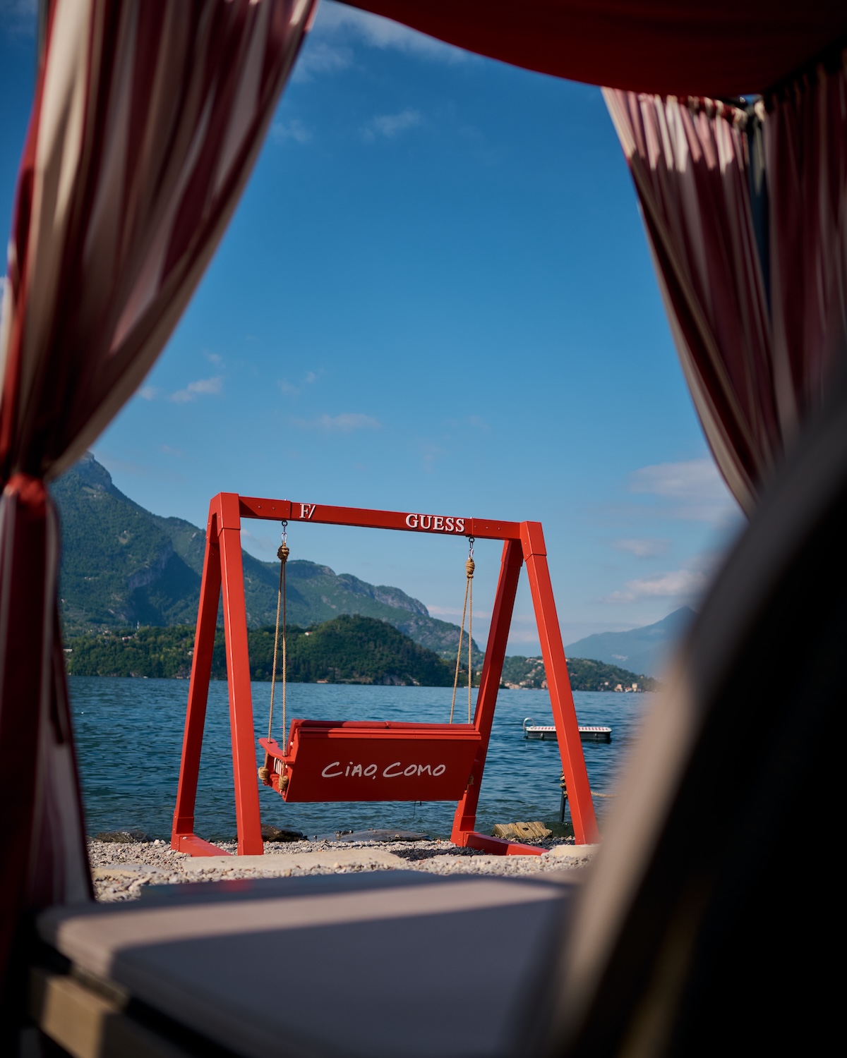 Red GUESS Ciao Como swing on Filario private beach, Lezzeno shoreline framed by striped curtains, Lake Como mountain backdrop, design hotel installation | TheAficionados.com