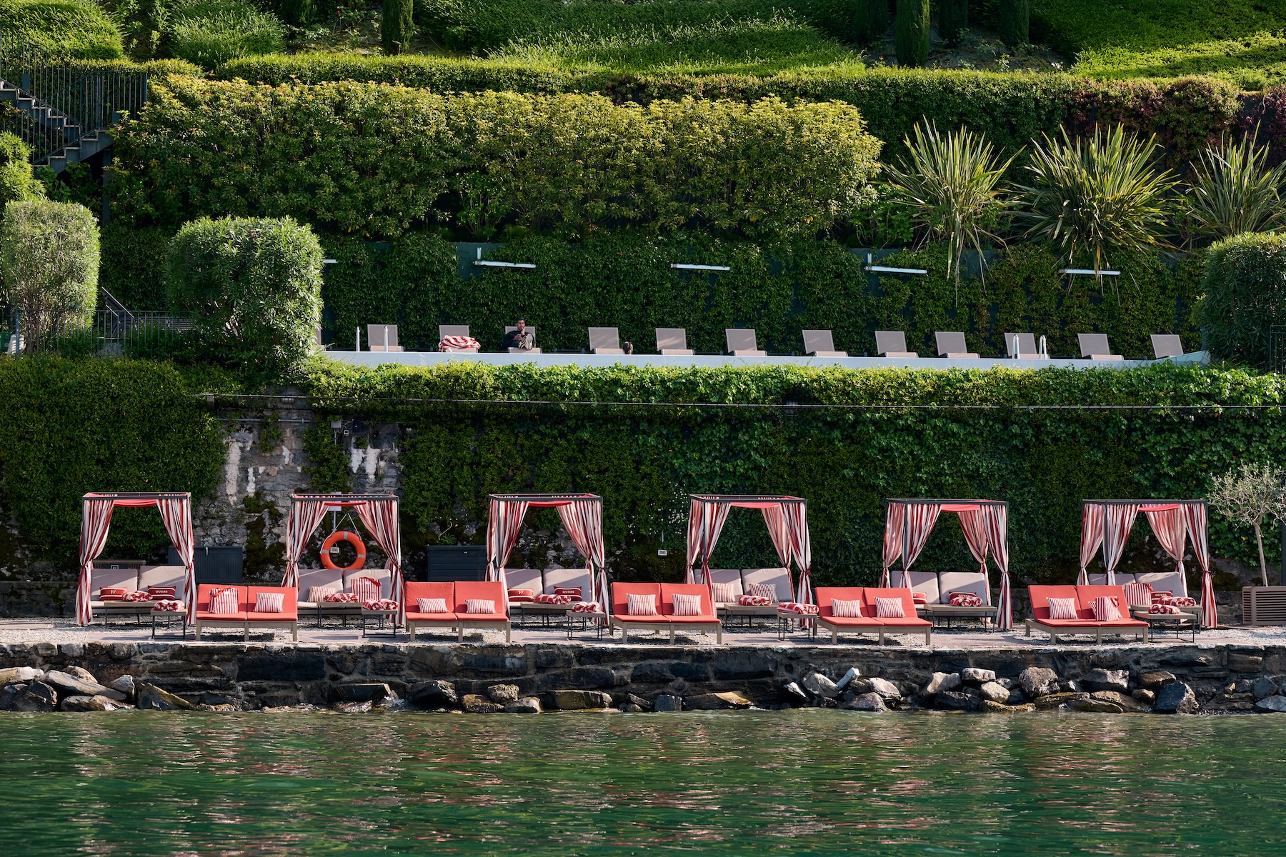FILARIO Hotel in partnership with GUESS | Row of red-and-white striped cabanas and loungers along a hedged waterfront on Lake Como