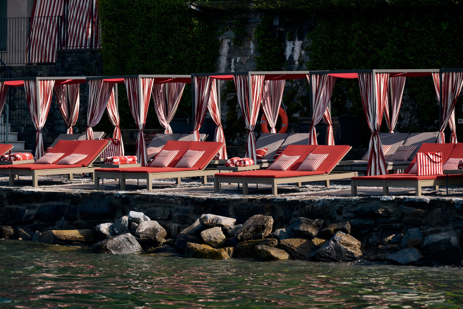 Row of red sun loungers with red-and-cream striped cabana curtains along Filario stone shoreline, GUESS Ciao Como beach club, Lake Como design hotel | TheAficionados.com