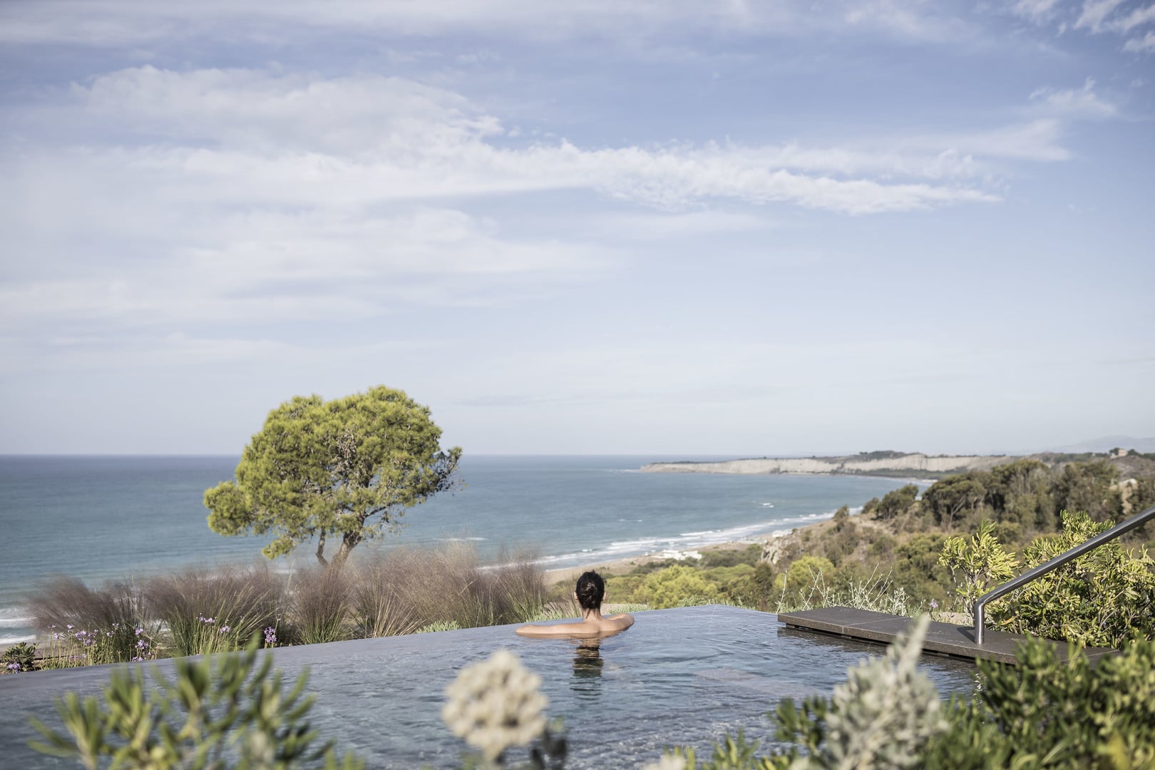 ADLER Sicilia Woman in a dark infinity pool gazing out over the Sicilian coast and a lone pine near Agrigento