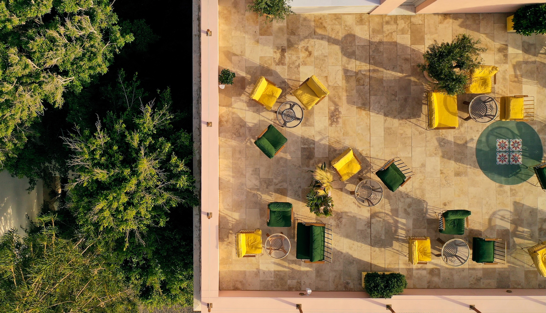 Overhead view of the courtyard terrace at Aristide Hotel, Syros, yellow and green cushioned seating on travertine paving shaded by mature trees, a neoclassical design hotel in Hermoupolis.